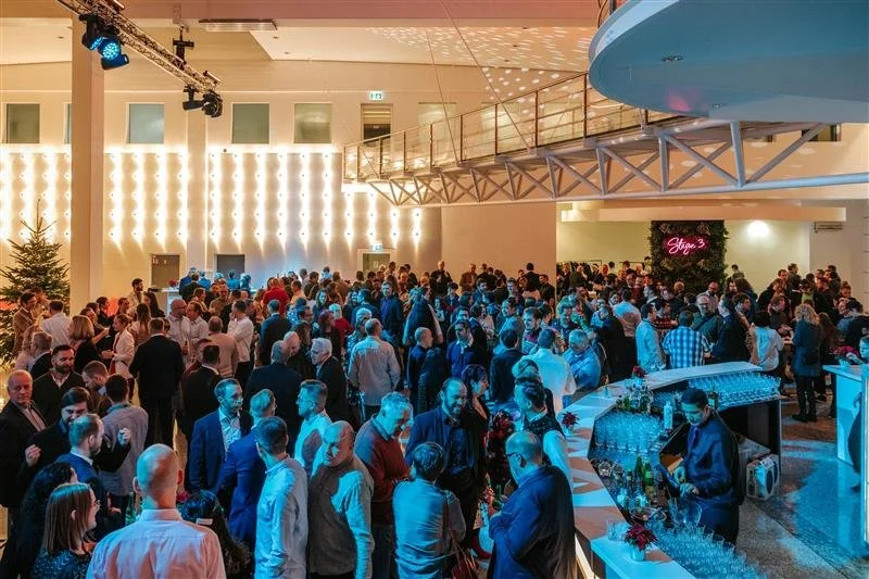 Menschen versammeln sich bei einer Party in einem großen Saal mit dekorativem Hintergrund und Bar, festliche Beleuchtung, Weihnachtsbaum, und einer Bühne.