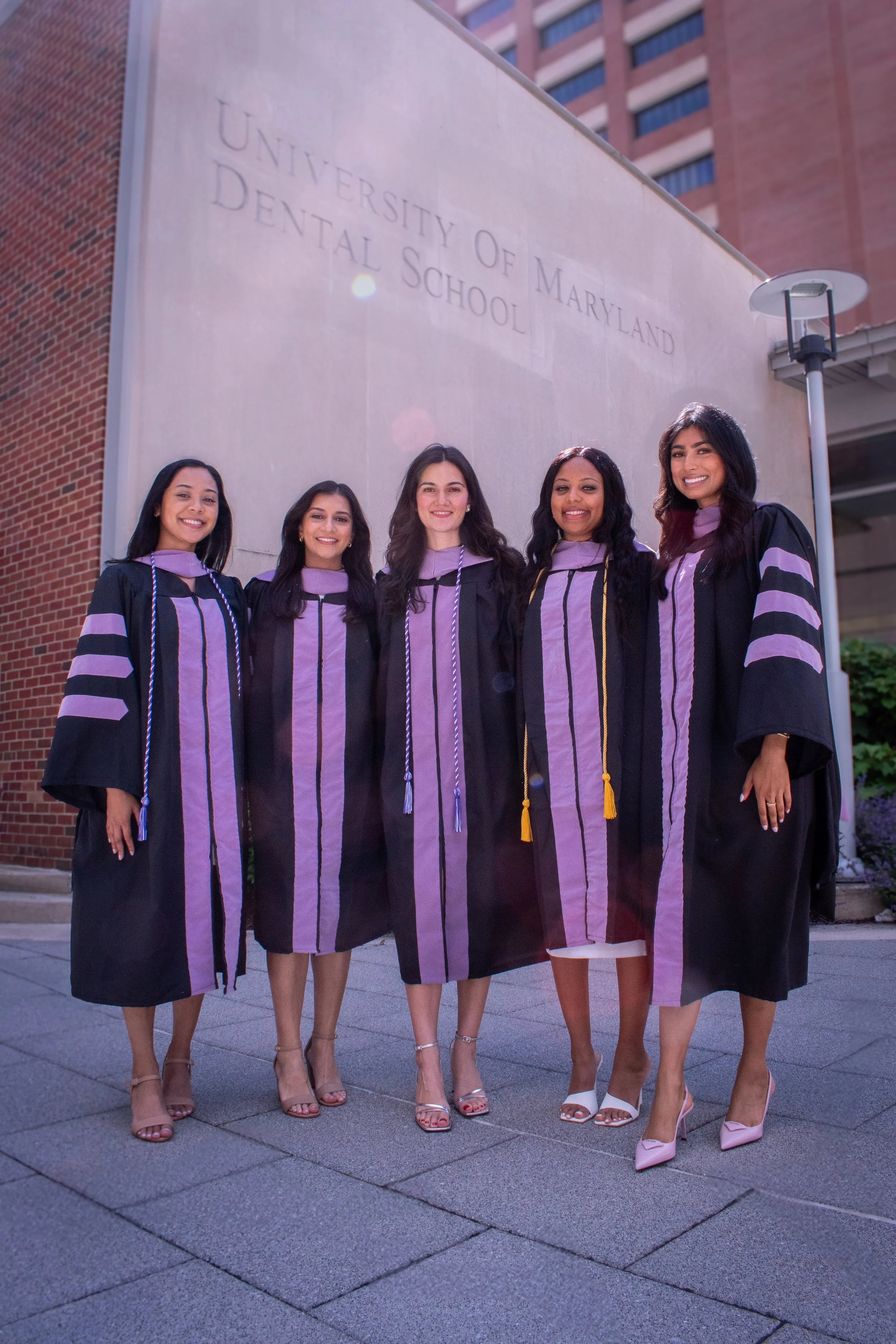 Five friends pose together in graduation regalia outside University of Maryland Dental School, beaming with pride. Purple-trimmed doctoral robes catch evening light as the newly minted dentists celebrate their shared journey and accomplishment.