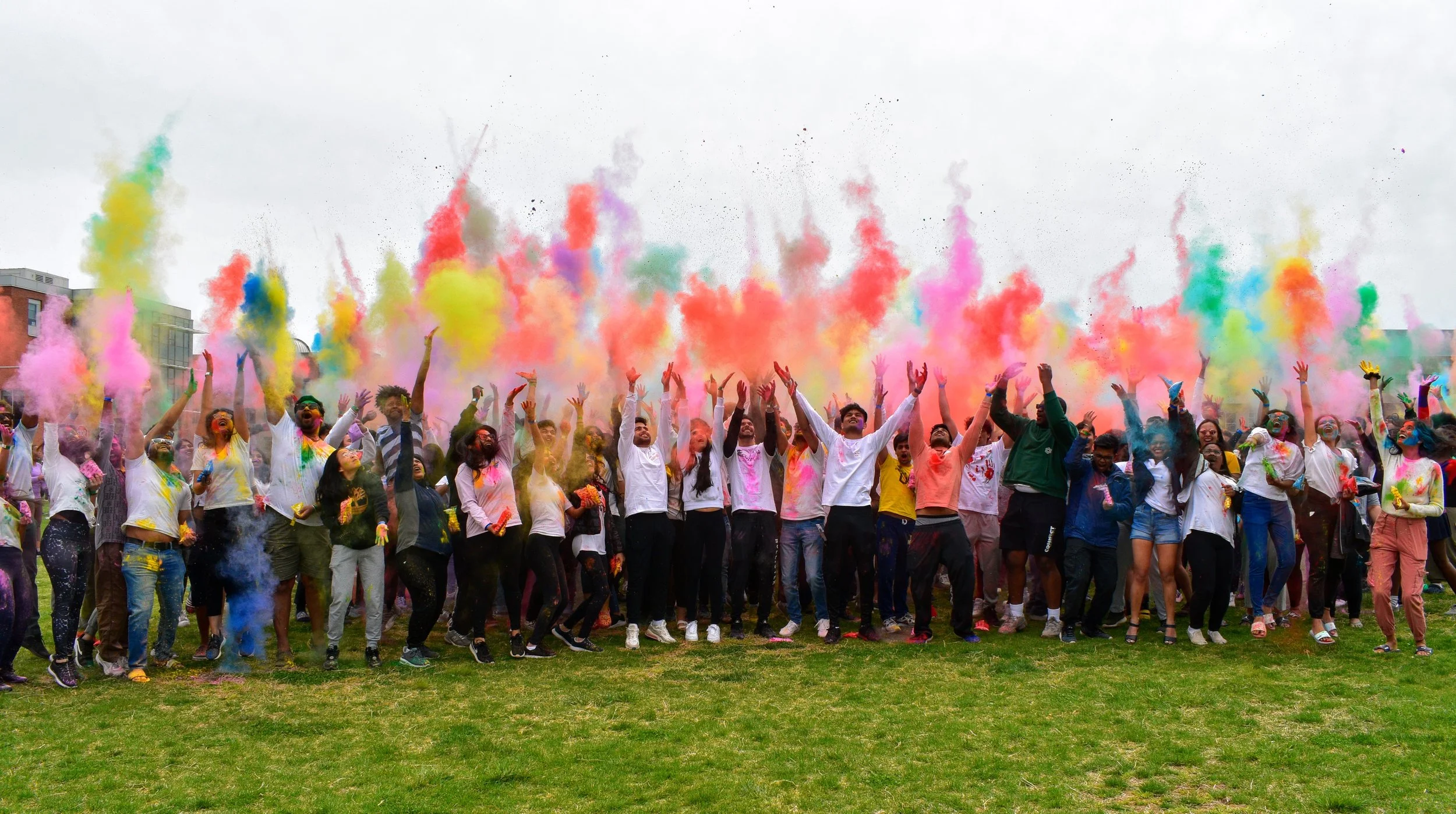 Large crowd outdoors celebrating with Holi powder, throwing bright clouds of pink, yellow, blue, green, and orange into the air, people in casual clothes cheering and jumping on a grassy field under an overcast sky.