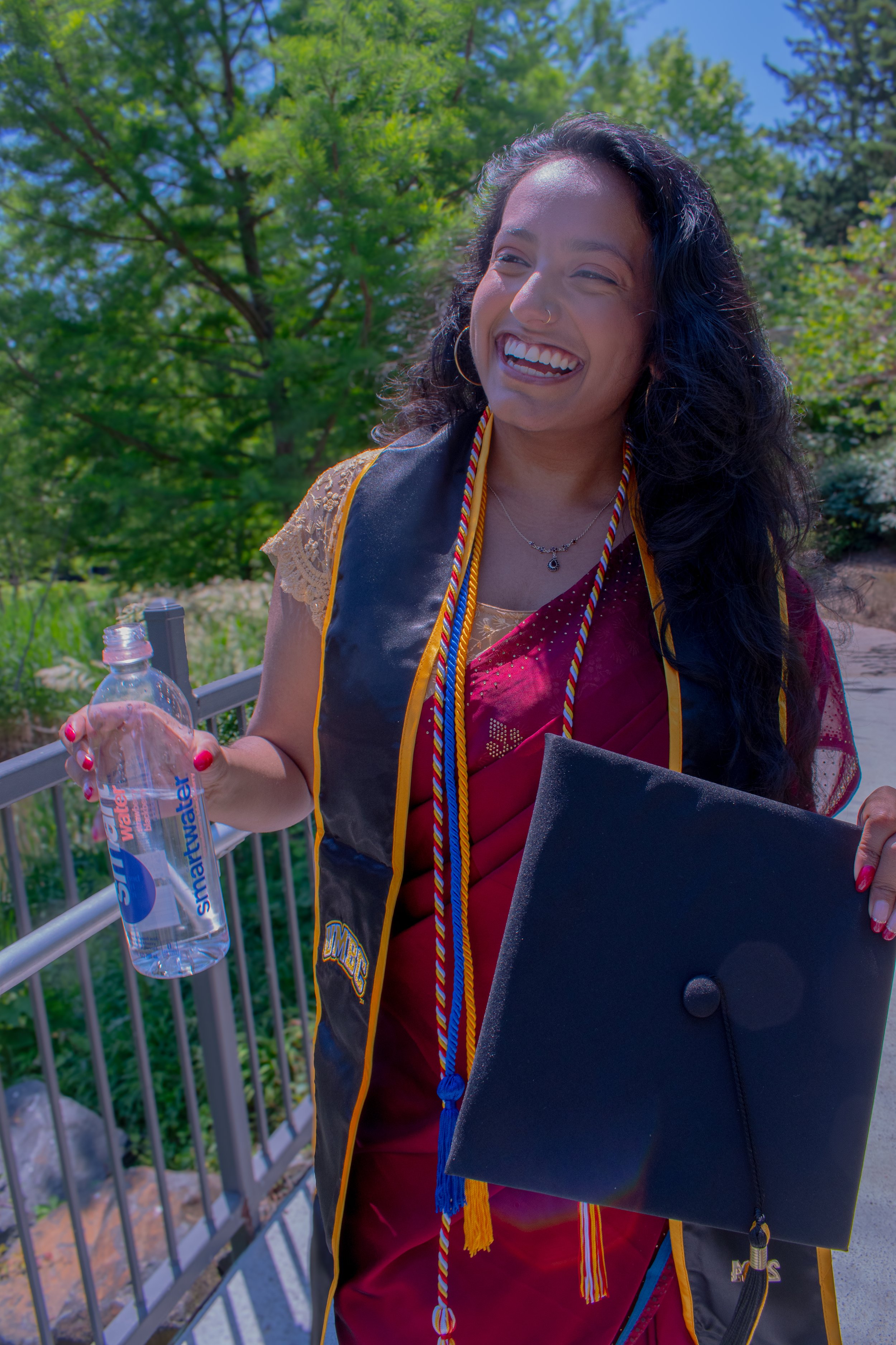 Graduate beams with joy in black regalia and honor cords, holding diploma and water bottle on sunny commencement day. Red embellished outfit peeks beneath graduation gown as she celebrates achievement against backdrop of vibrant greenery