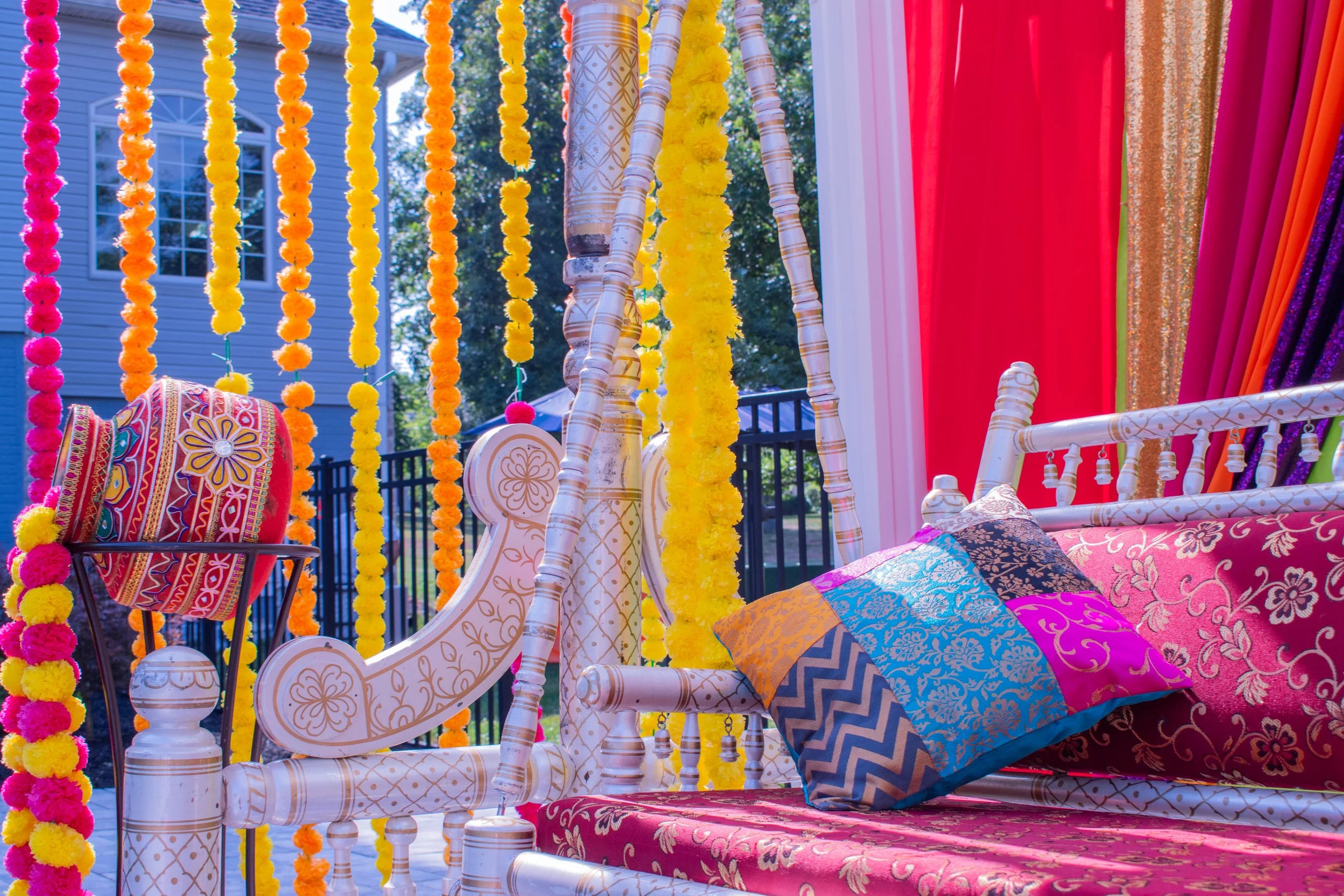 Ornate carved daybed draped with jewel-toned pillows sits beneath vibrant marigold garlands in pink, orange, and yellow. Rainbow curtains frame the mehndi celebration space, mixing traditional embroidered textiles with bold modern colors