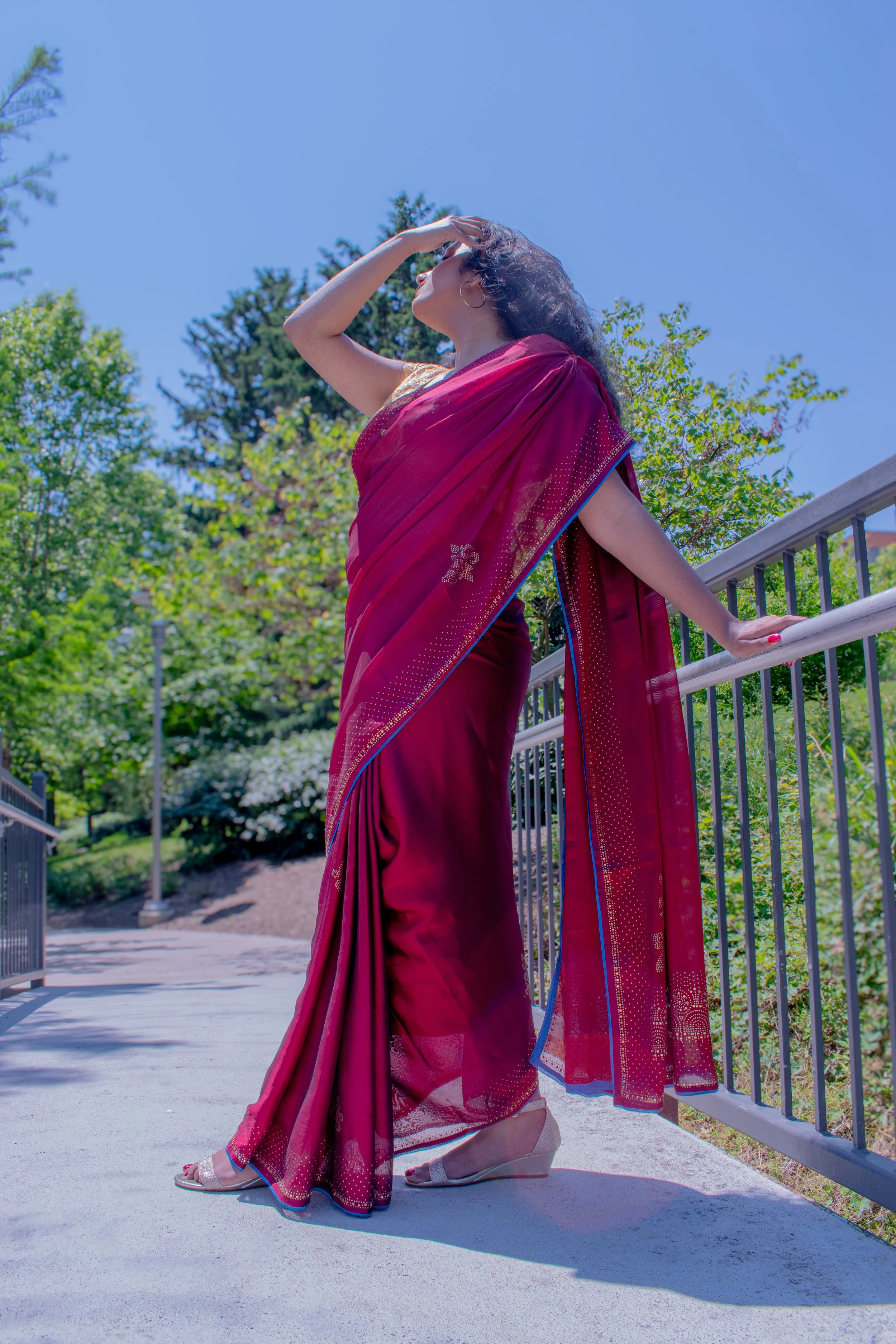 A woman in a red saree standing on a bridge outdoors, touching her forehead with her left hand, against a backdrop of trees and blue sky.