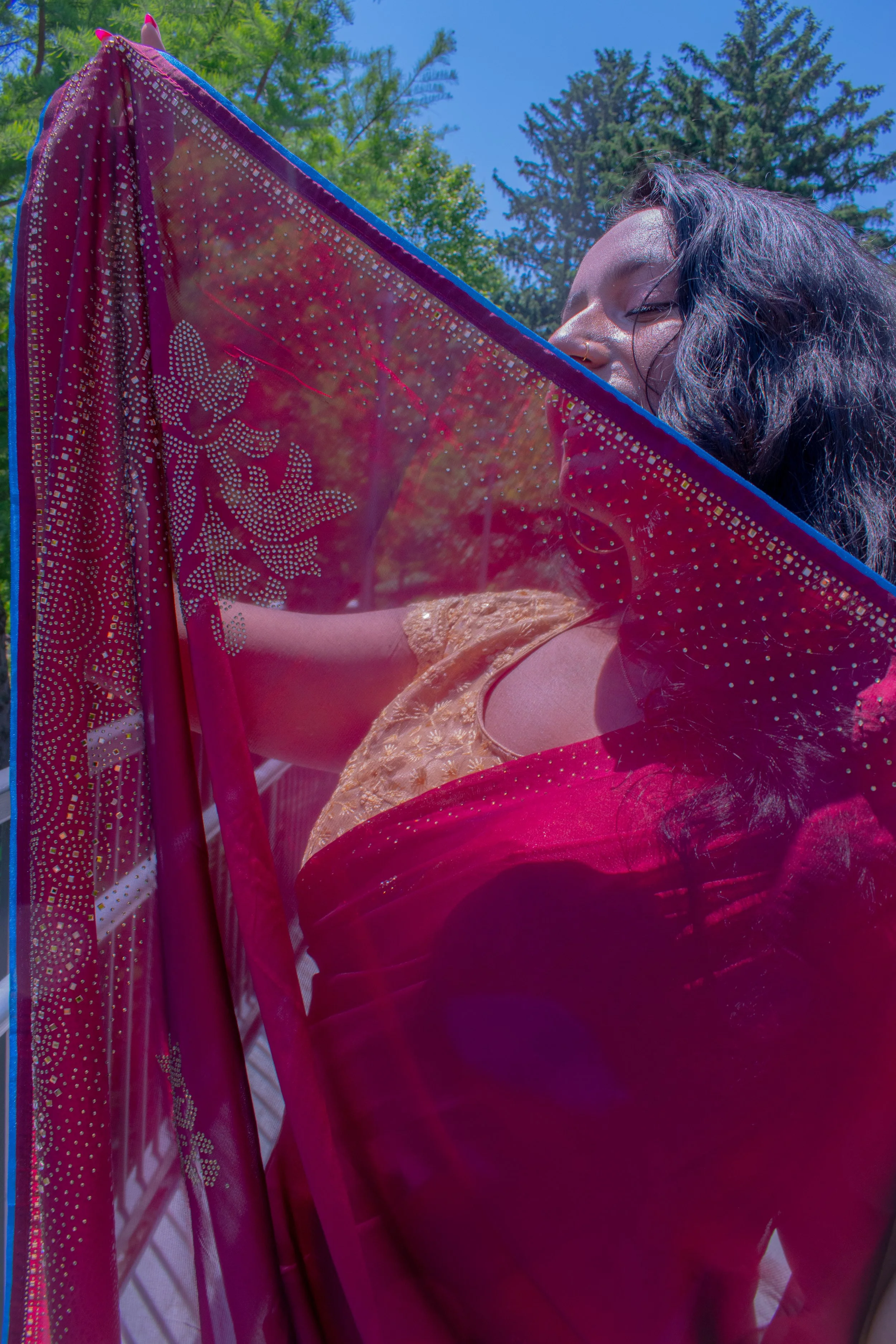 She peeks playfully from behind ornate red dupatta studded with crystals and golden embellishments, sunlight filtering through sheer fabric. Traditional South Asian bridal attire glows against lush green trees and blue summer sky