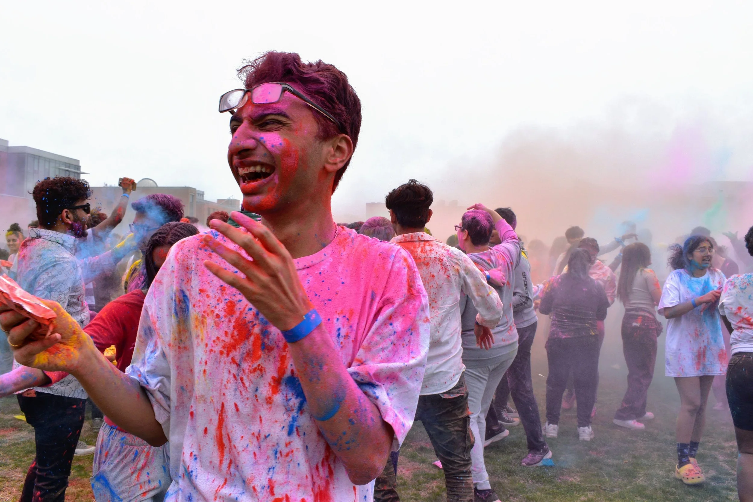 Holi. A student laughs with their mouth open, covered in bright pink, blue, and red powder, holding a packet of color. Around them, a crowd dances and throws powder into the air, filling the field with colorful clouds and a lively festival vibe