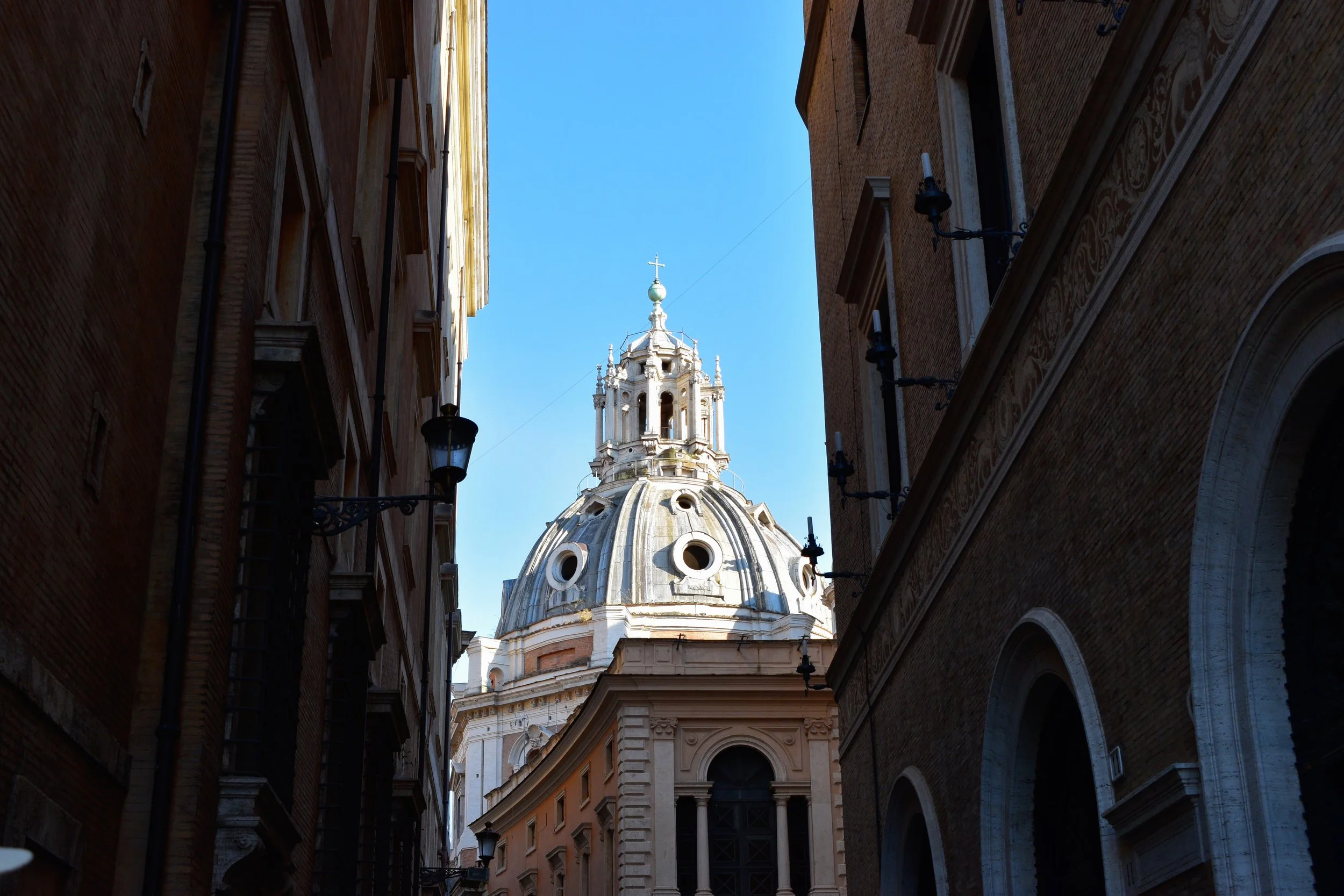 A sunlit church dome rises between narrow brick buildings, framed by old streetlamps, with pale stone details glowing against a clear blue sky in a quiet Roman street.