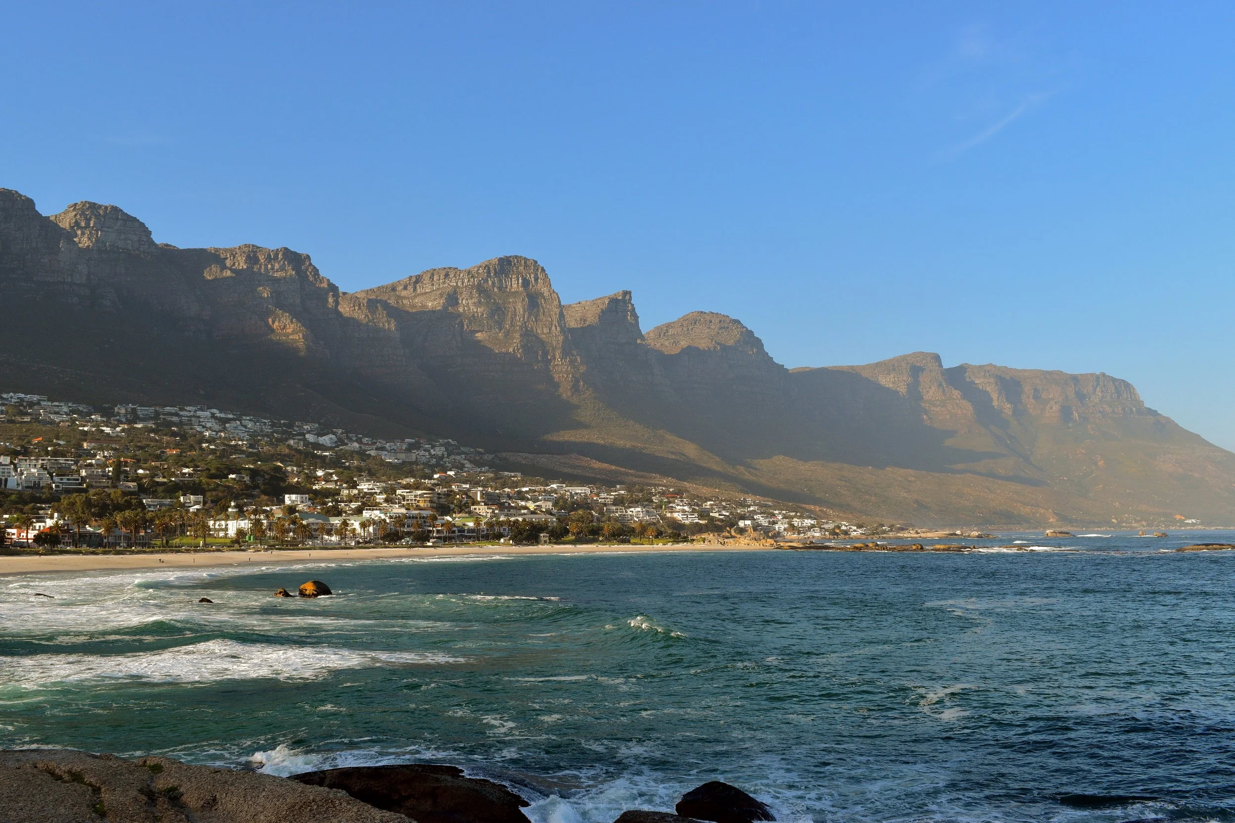 Cape Town: Coastal town at the foot of rugged mountains, white buildings lining a sandy beach as waves roll in under a clear blue sky at golden hour.