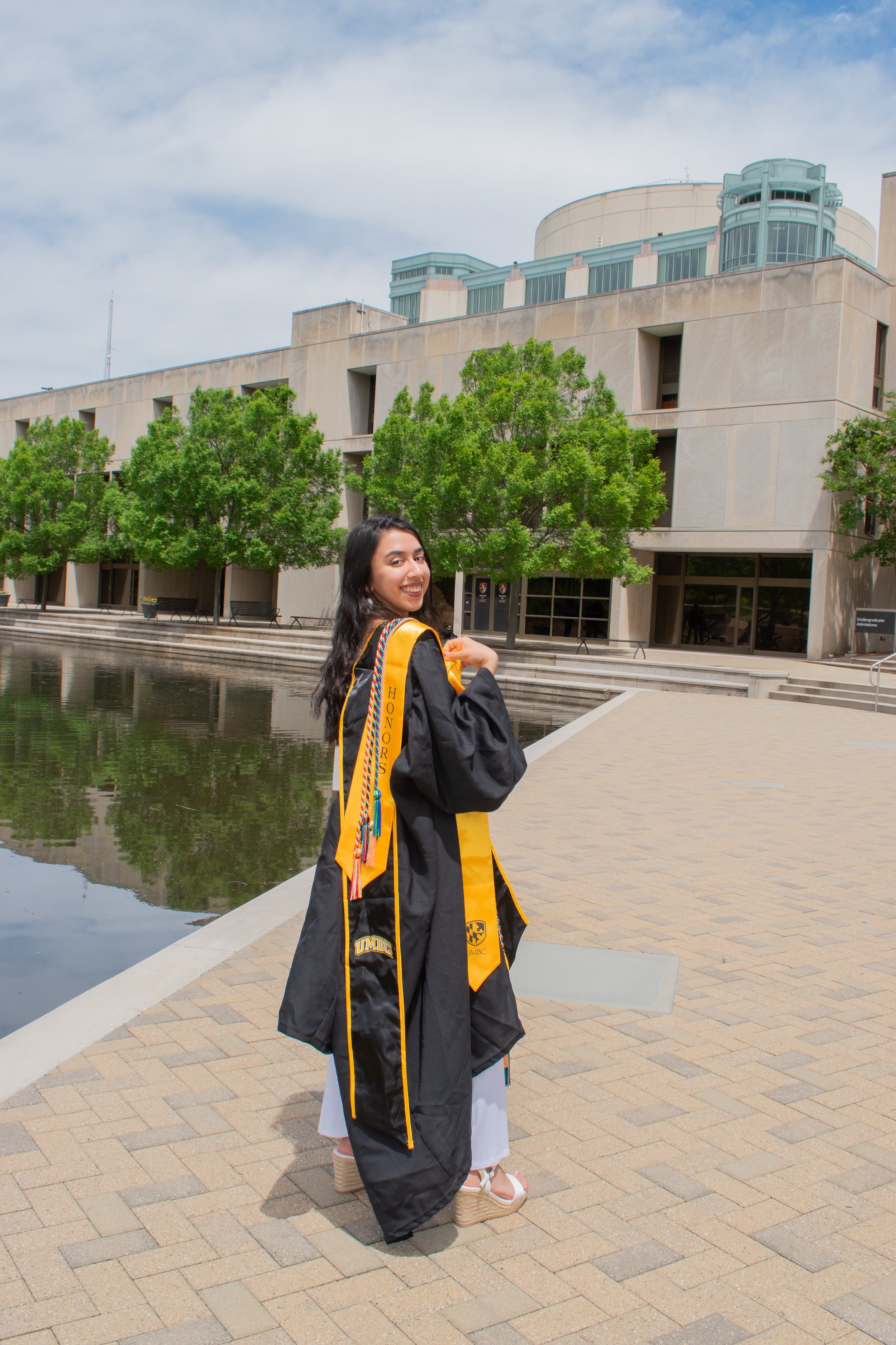 A happy Mashaal looks back at the camera, wearing her black UMBC graduation regalia, cords, and a gold Honors stole. In the background is a pond, trees, and UMBC's iconic library
