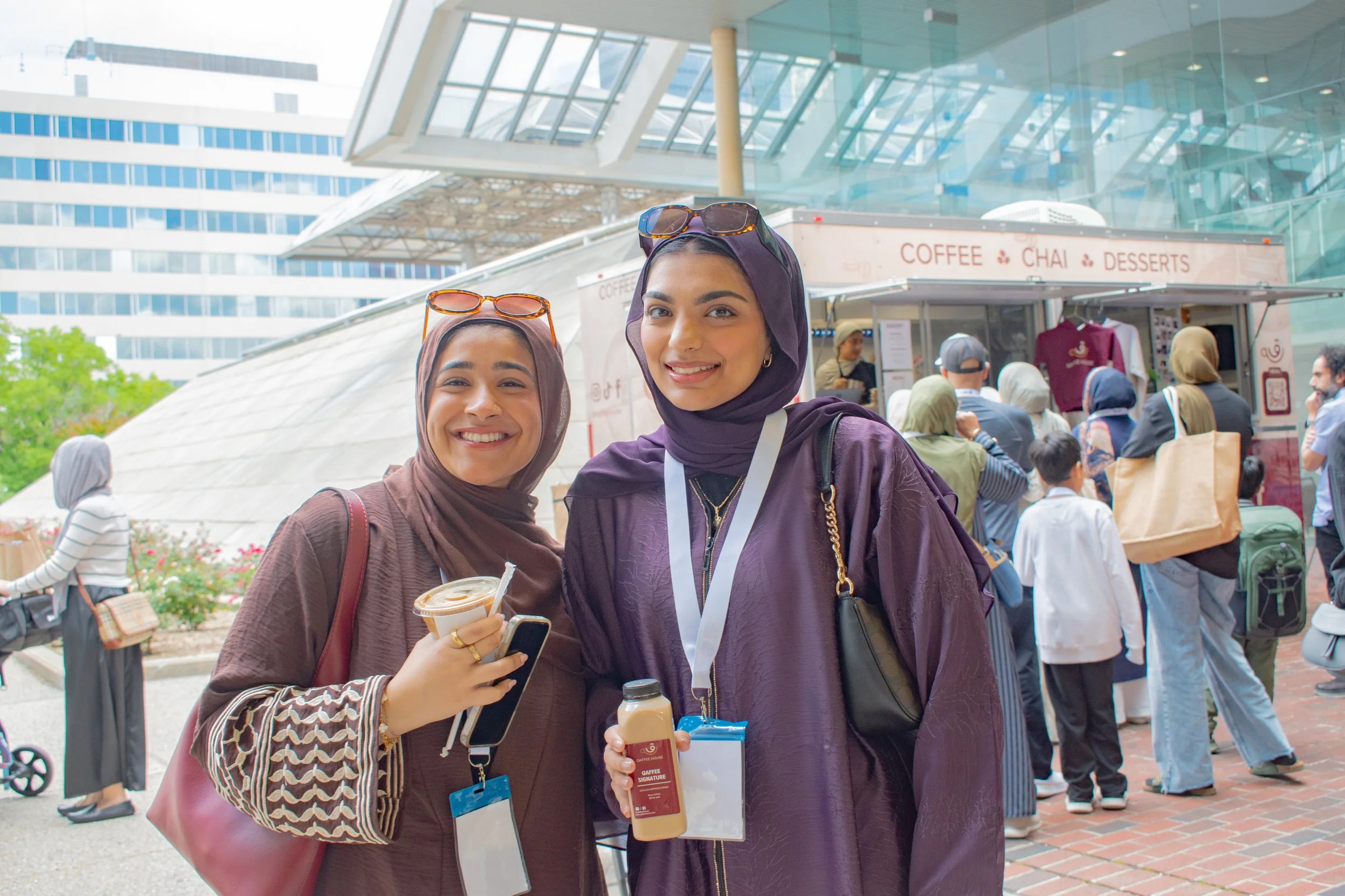 Two friends pose with iced drinks outside café truck at sunny gathering, both wearing purple hijabs and sunglasses. Modern building with dramatic angles frames the scene as community members mingle around coffee and chai vendor.