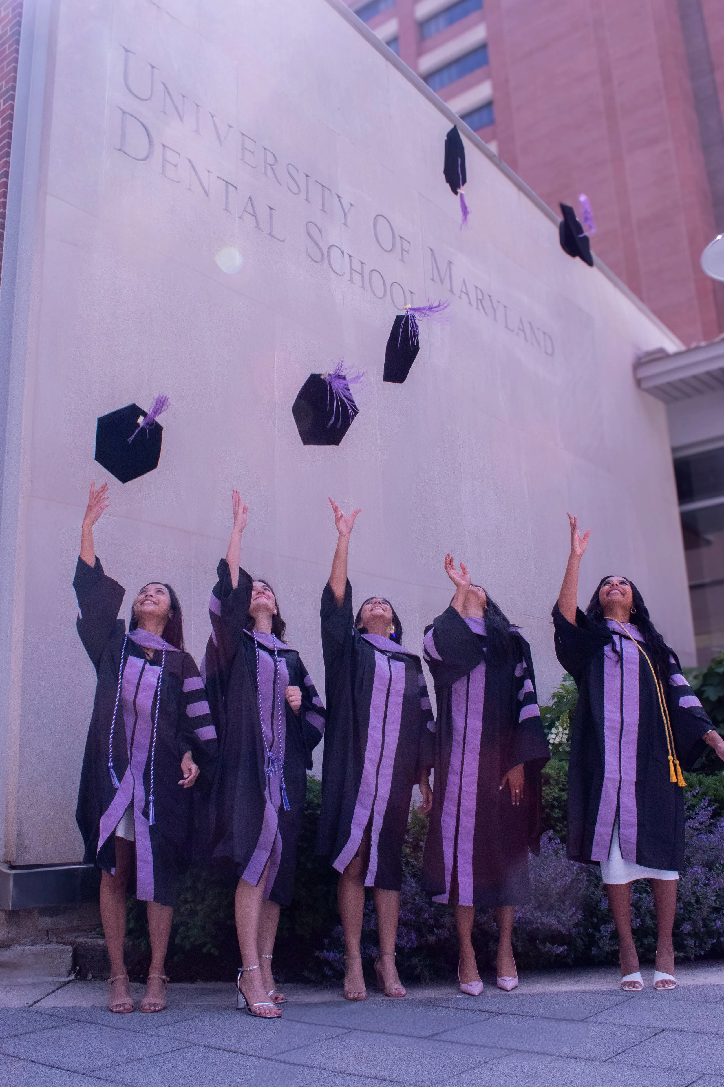 Five dental school graduates toss their caps skyward in synchronized celebration beneath University of Maryland Dental School sign. Black robes with purple trim flutter as the new dentists mark their achievement at golden hour on campus.