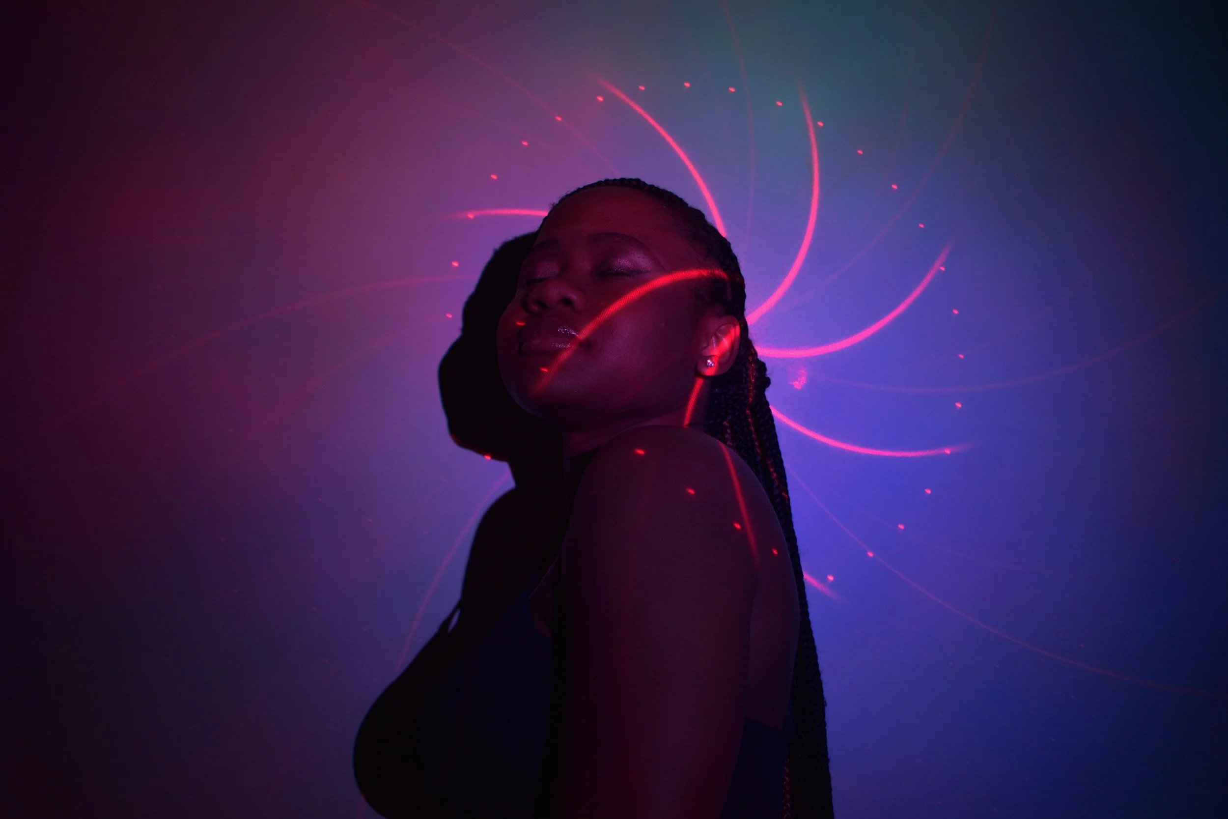 Alberta  in profile against a dark wall, lit by pink and blue neon-style light with swirling laser patterns forming a halo-like glow behind their head.