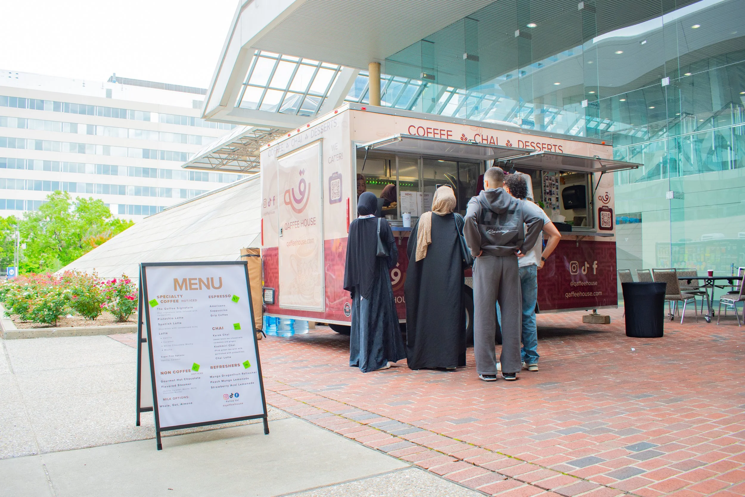 Customers line up at coffee and chai food truck parked outside modern building, menu board displayed prominently. Glass architecture soars above brick plaza where community members gather for specialty drinks and desserts on pleasant day
