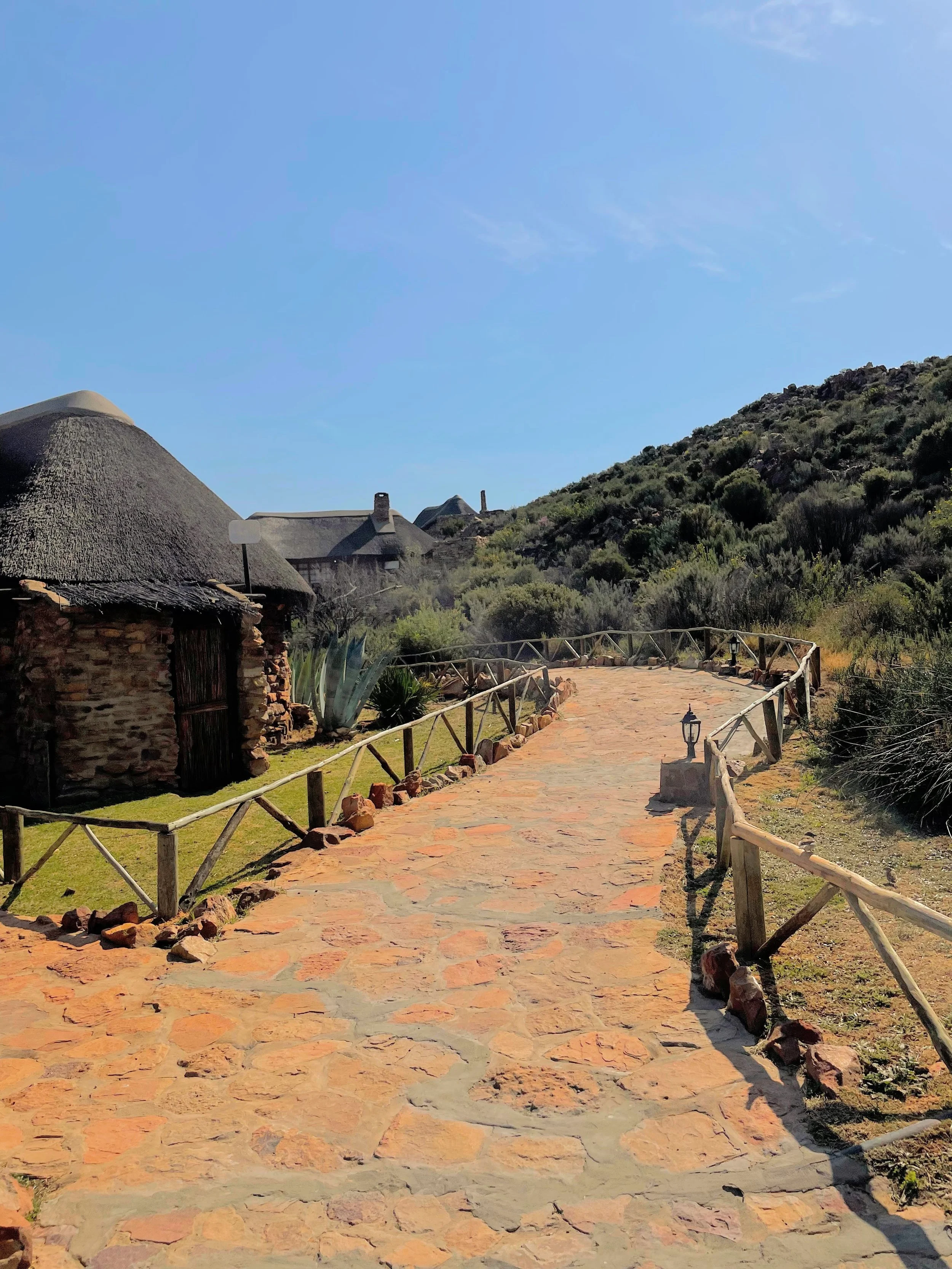 A stone pathway leading through a rustic, village-like area with thatched-roof houses and green hills in the background, under a clear blue sky.