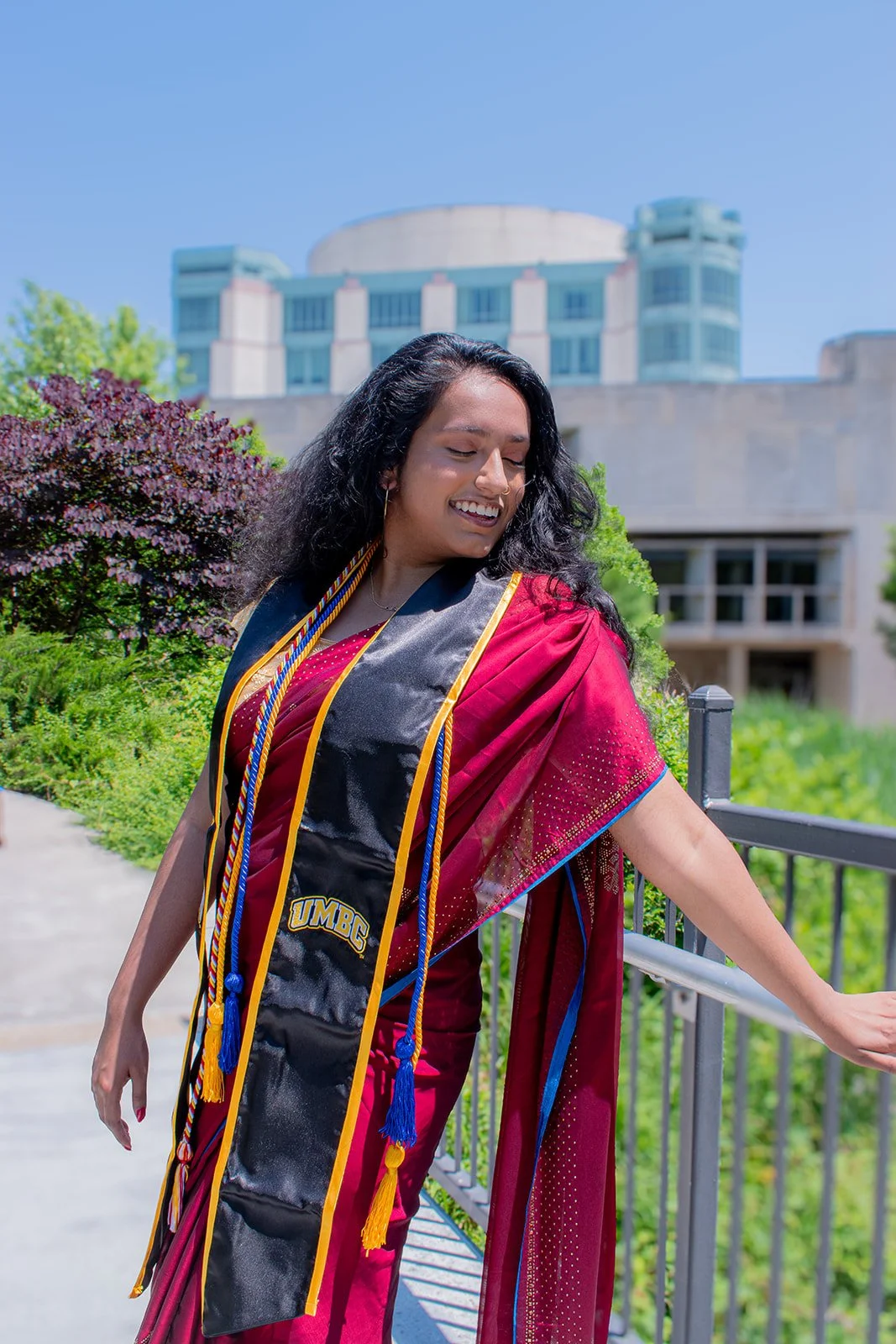 Sivalesh wears a vibrant red and gold sari with black graduation stole and UMBC cords smiles joyfully on campus walkway with modern university building in background.