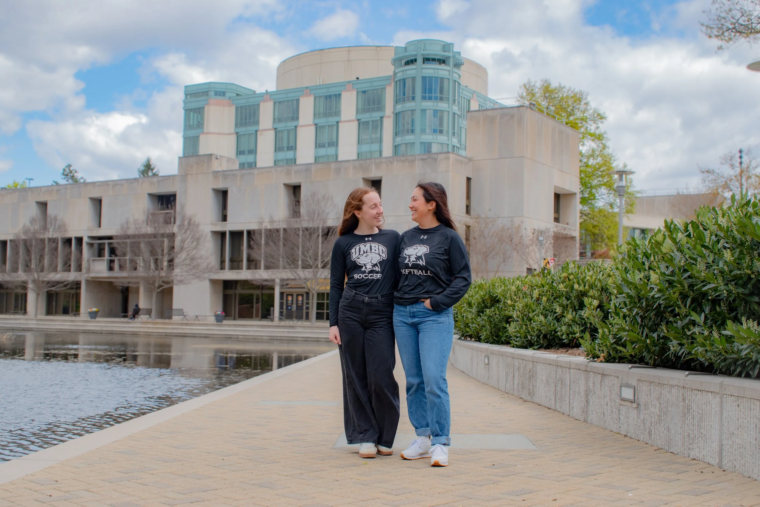 Two friends walk side by side on campus pathway, sharing a laugh beside reflecting pool. Modern university buildings with blue-green glass rise behind them as they wear matching athletic shirts, capturing an easy moment of college friendship