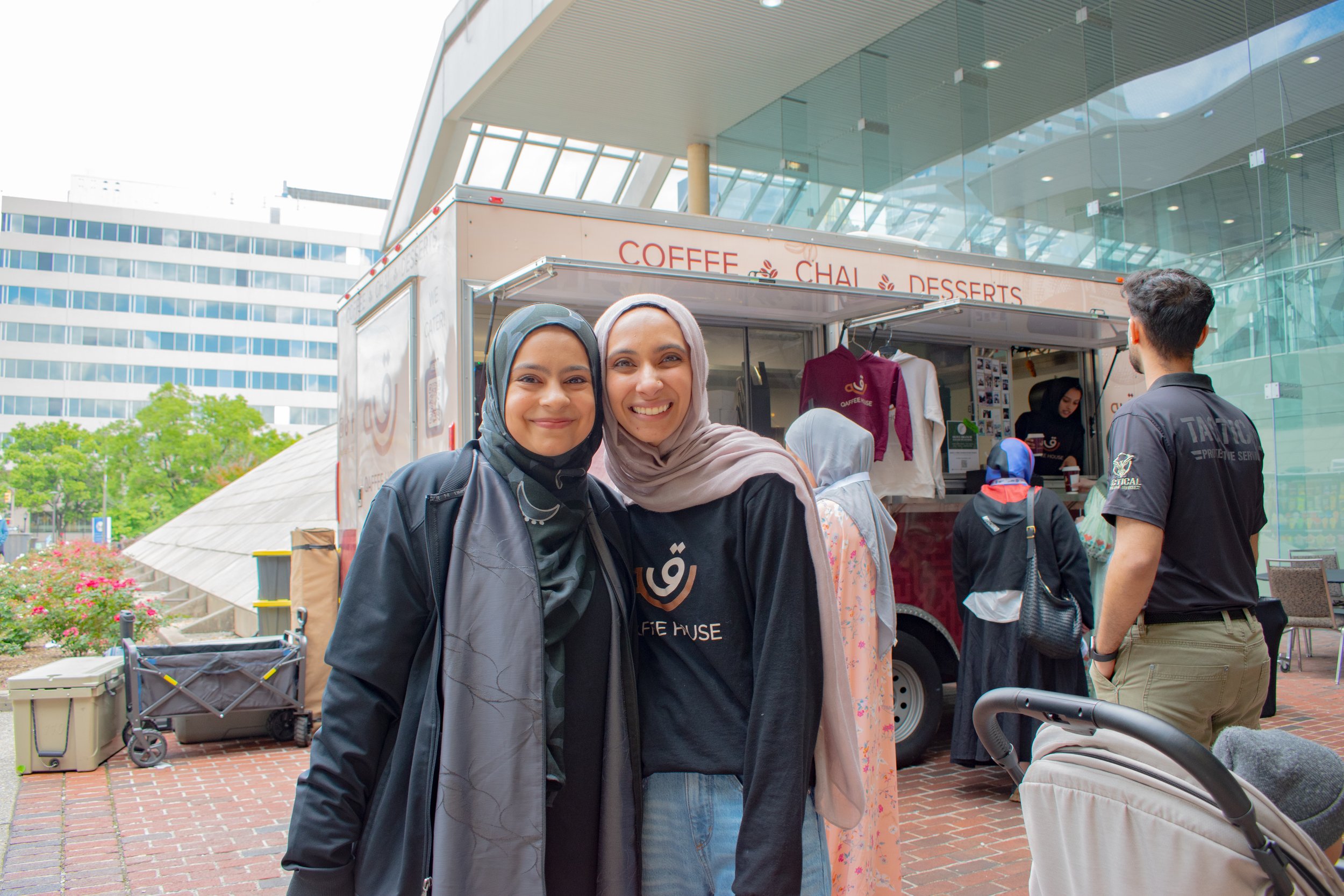 Friends smile together at community event, café truck visible behind them. Younger woman wears dark hijab while older woman in taupe hijab sports café branded shirt, capturing warm moment at outdoor gathering