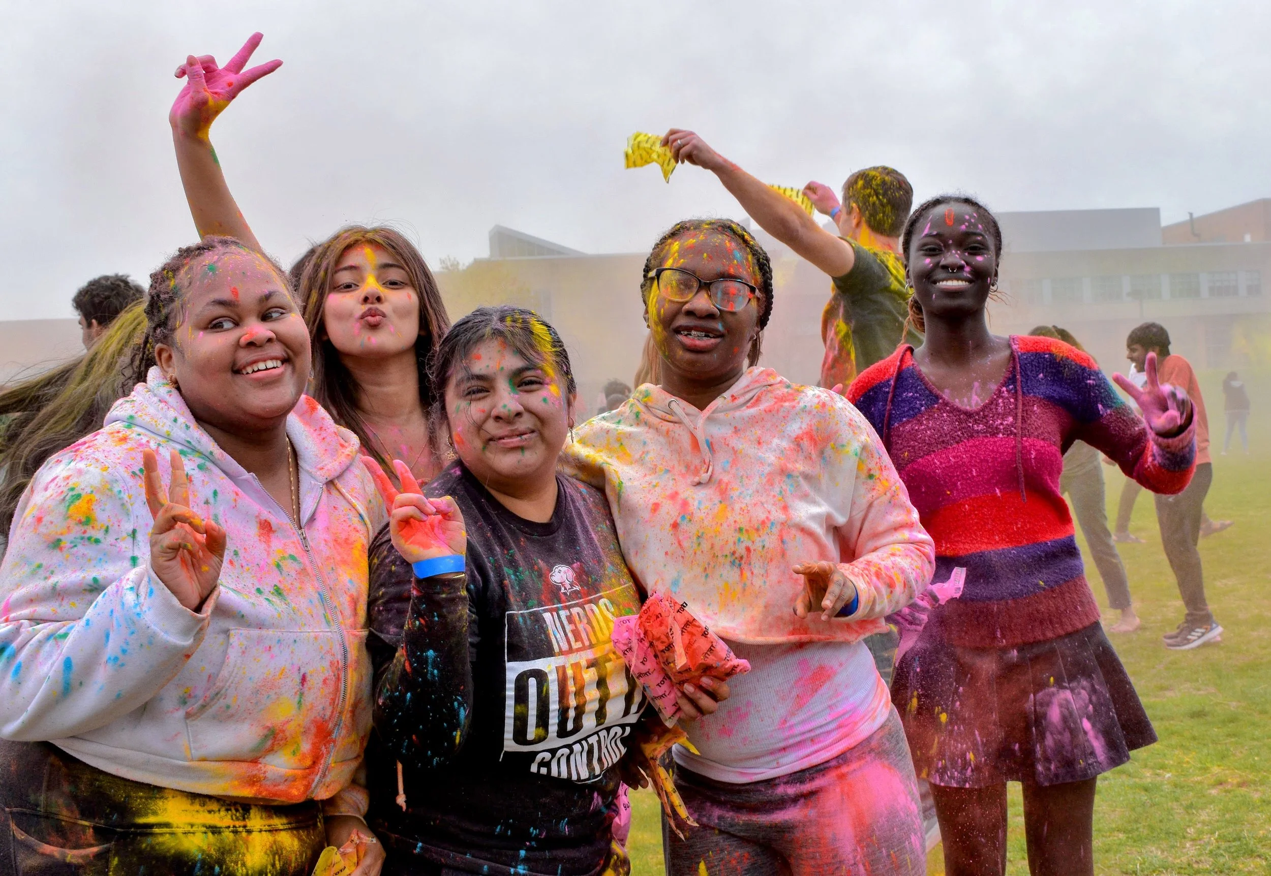Holi. Five friends stand together on a grassy field, smiling and throwing peace signs, covered head to toe in bright colored powder. The air is filled with color as others celebrate behind them, creating a joyful, energetic festival atmosphere.