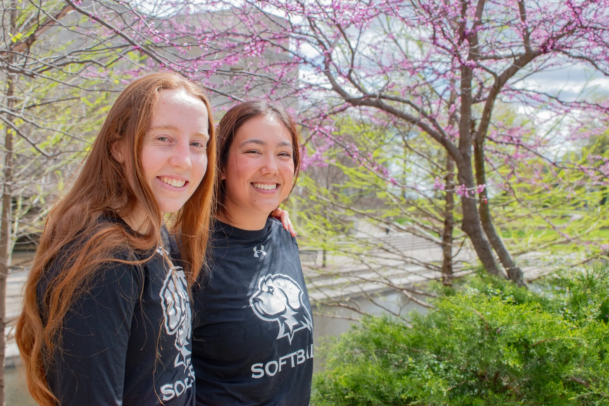 Two smiling female softball players in black jerseys with a lion logo, standing outdoors during spring with blooming pink cherry blossom trees in the background.