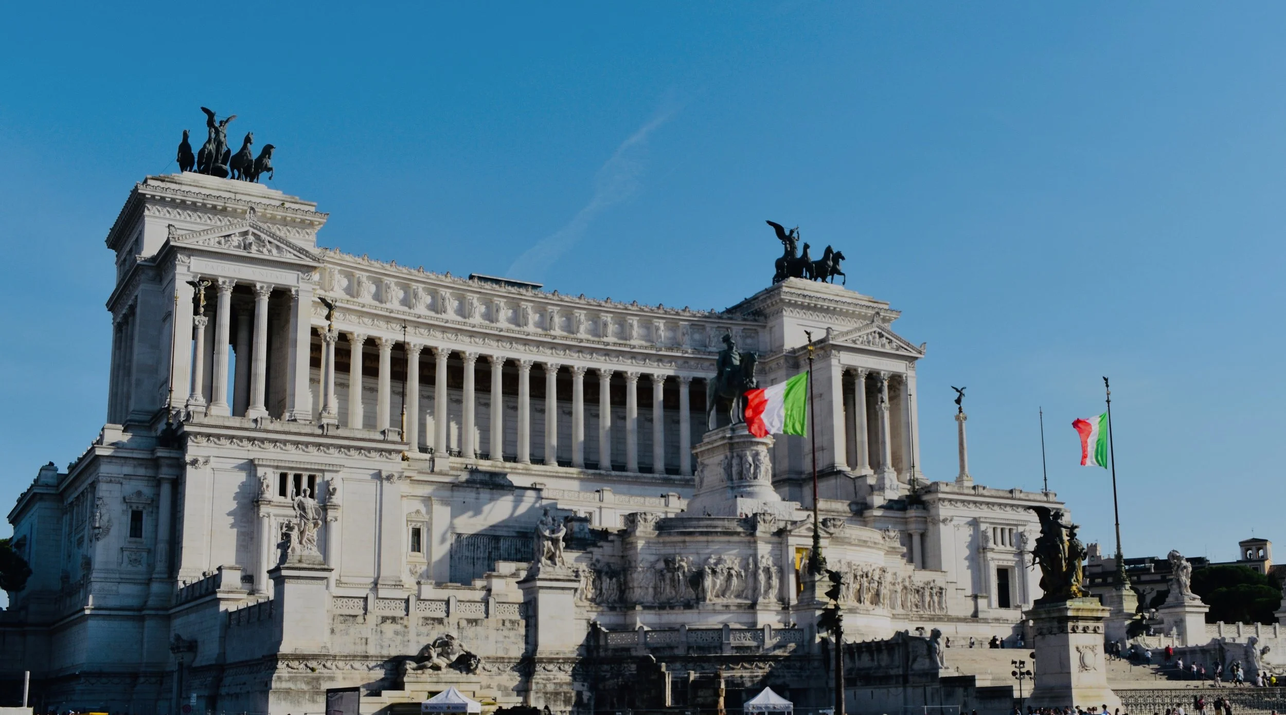 The Vittoriano monument in Rome gleams in white marble under a bright blue sky, with Italian flags waving and statues lining its grand terraces and columns.
