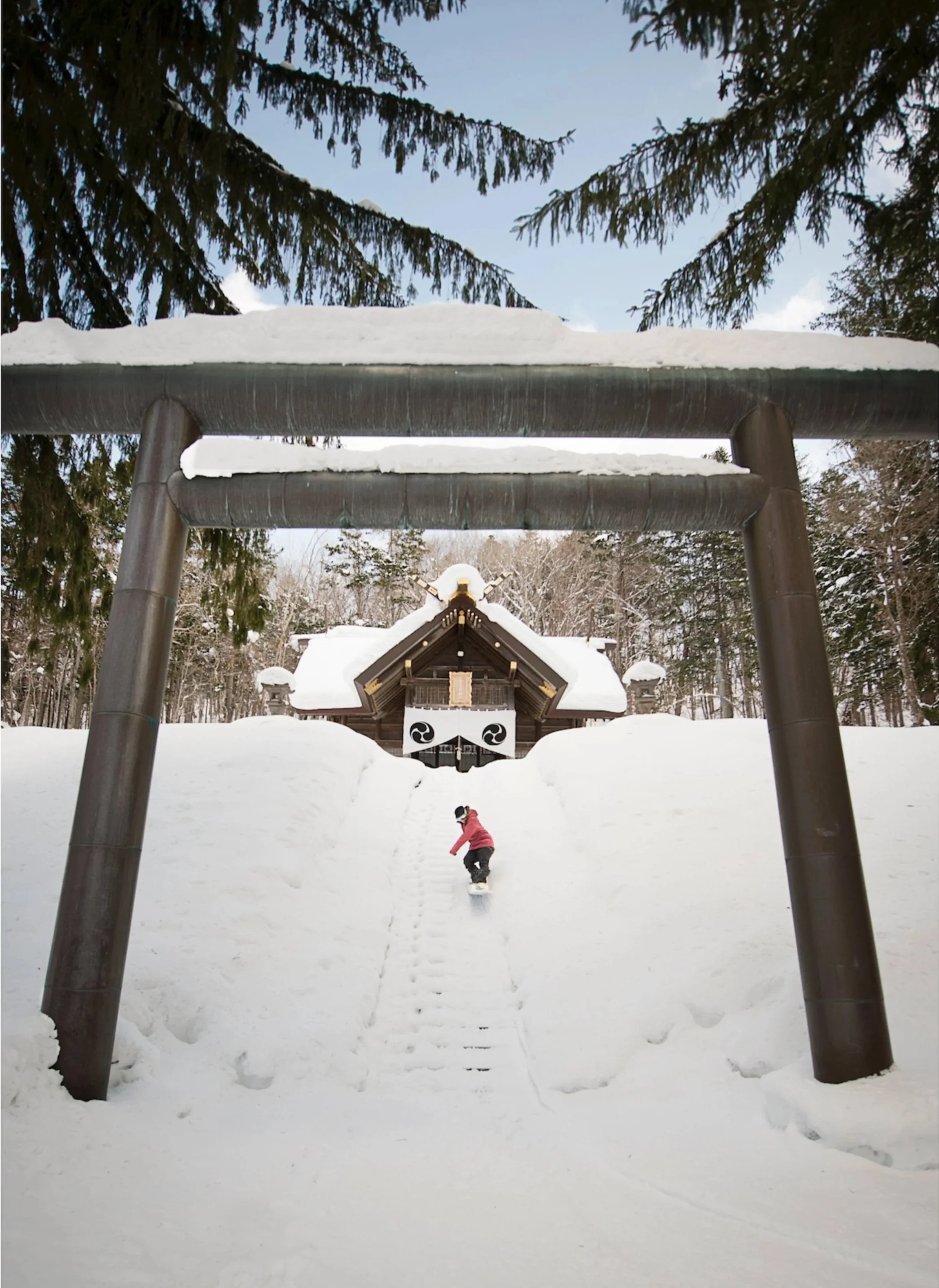 Une scène hivernale avec de la neige, un garçon en manteau rose en train de faire du snowboard spirituel en descendant une pente enneigée devant un temple japonais et un grand torii en bois dans un forêt enneigée.