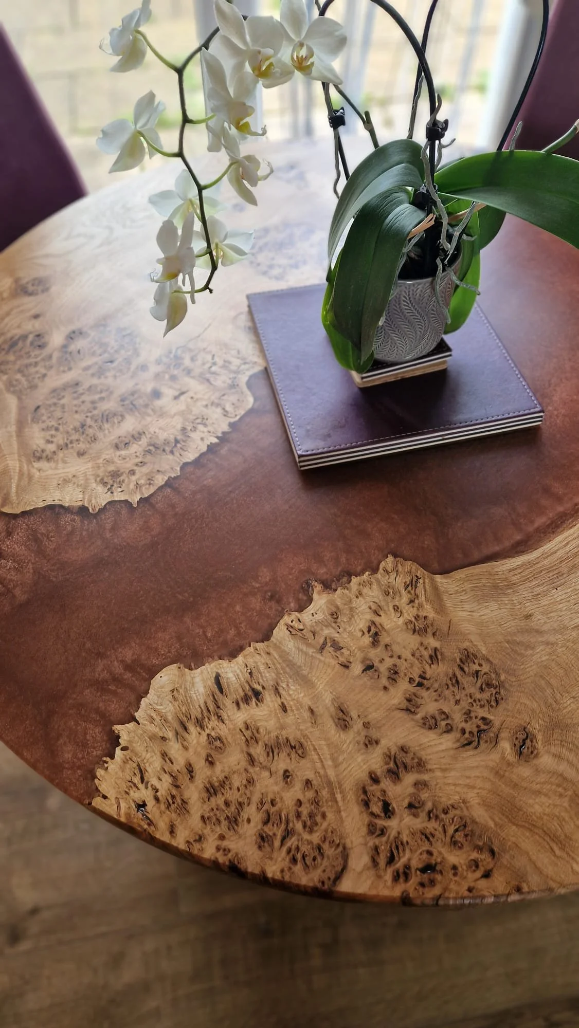 Close-up of a wooden table with natural grain patterns and a potted orchid flower arrangement on top, placed on a purple coaster and a stack of black notebooks near a window.