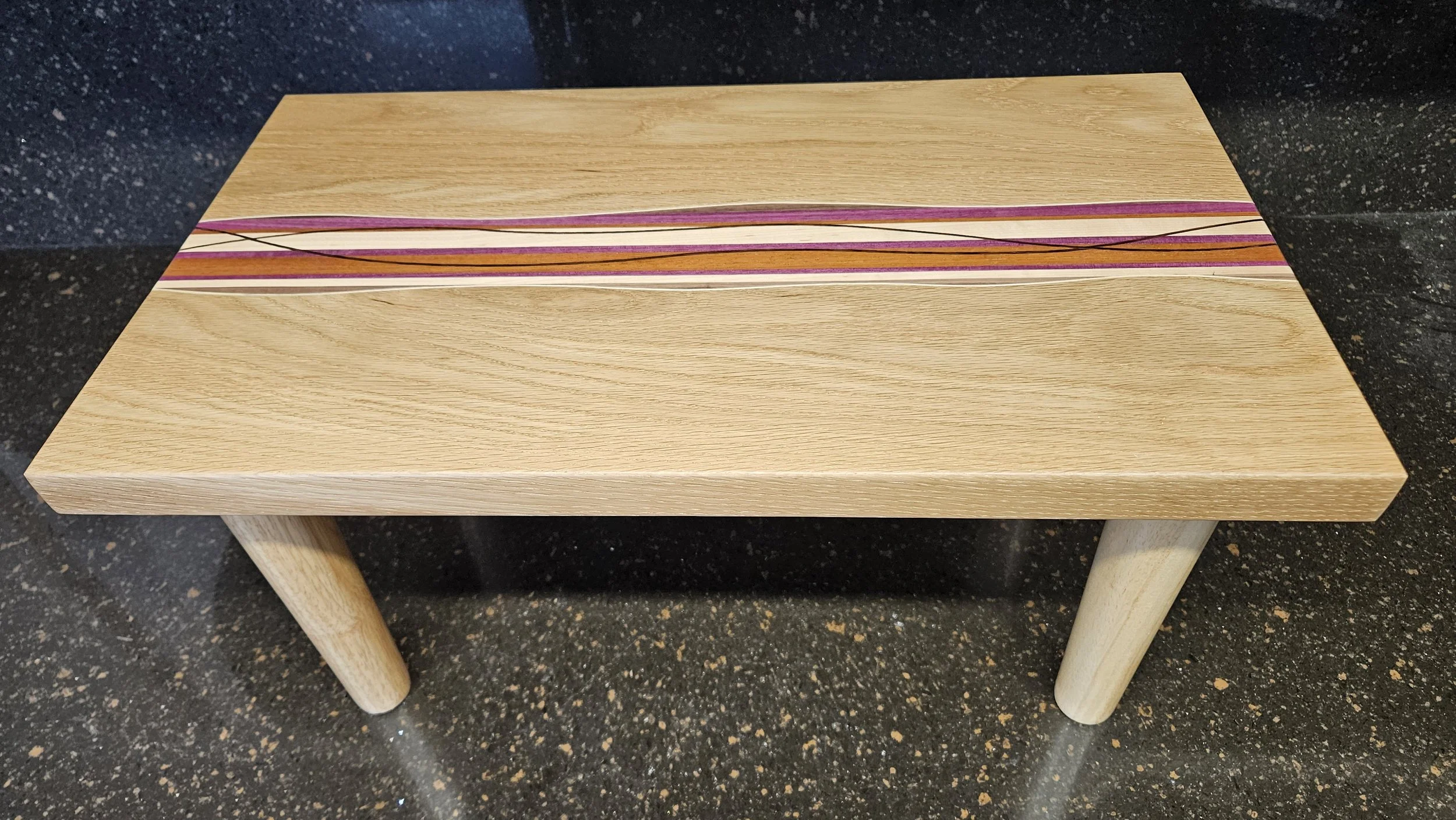 A rectangular wooden table with a colorful, striped inlay on top, positioned on a black speckled floor.