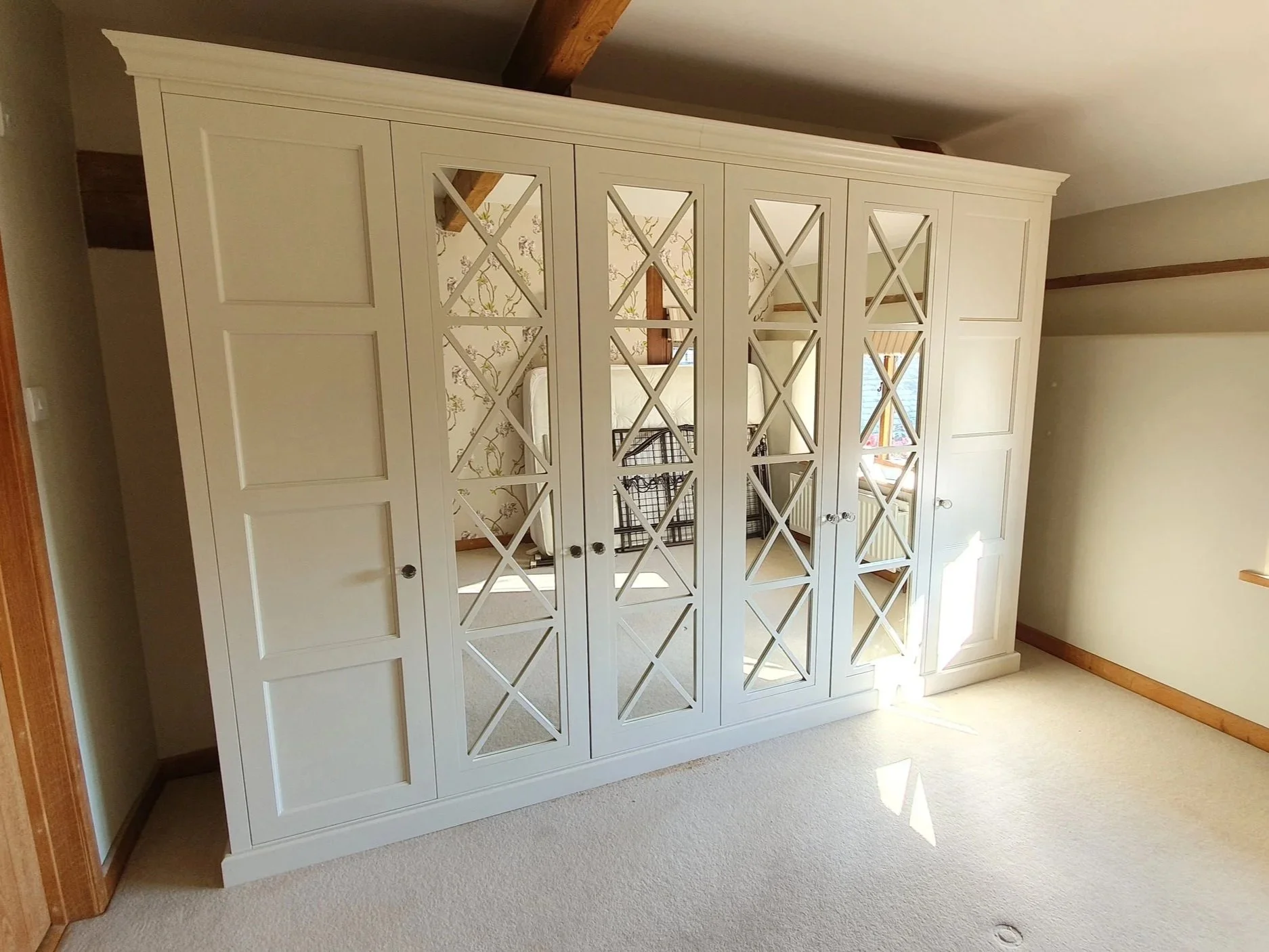 A white wardrobe with mirrored and paneled doors standing against a wall in a room with beige carpet and cream walls. Sunlight streams through a window, casting shadows on the floor.