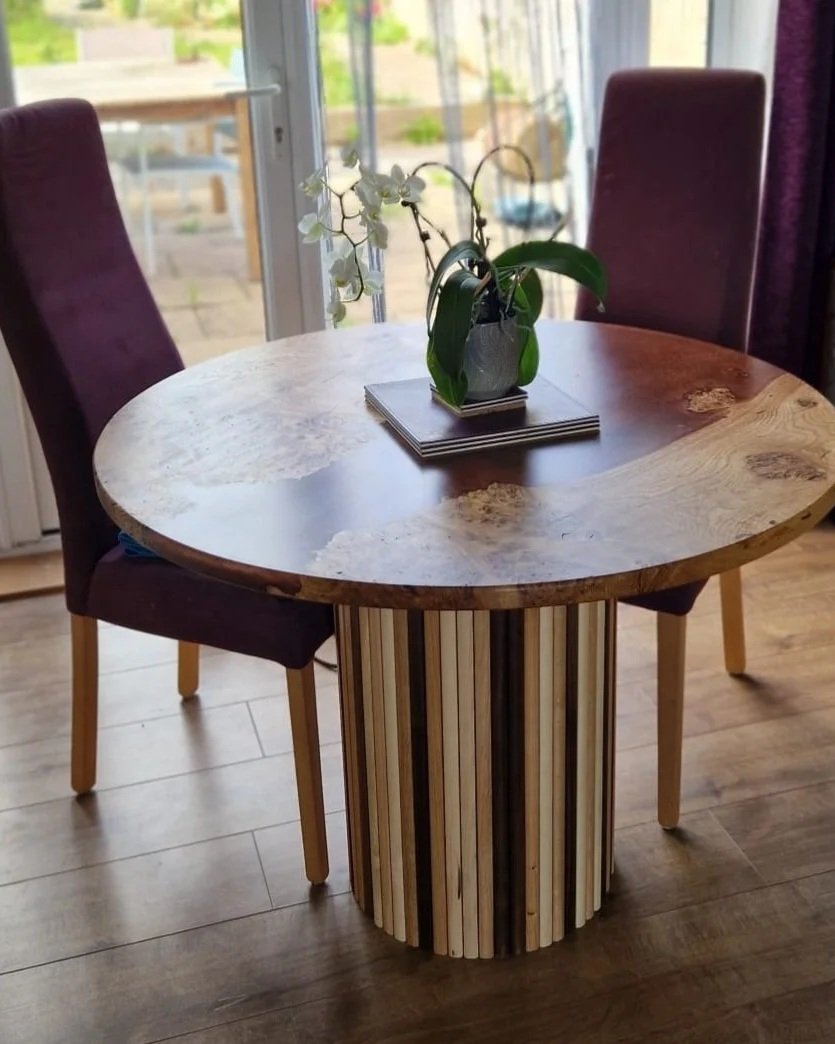 A round wooden dining table with a striped cylindrical base, three purple upholstered chairs, and a small vase with white orchids on top, situated in front of a sliding glass door leading to a patio