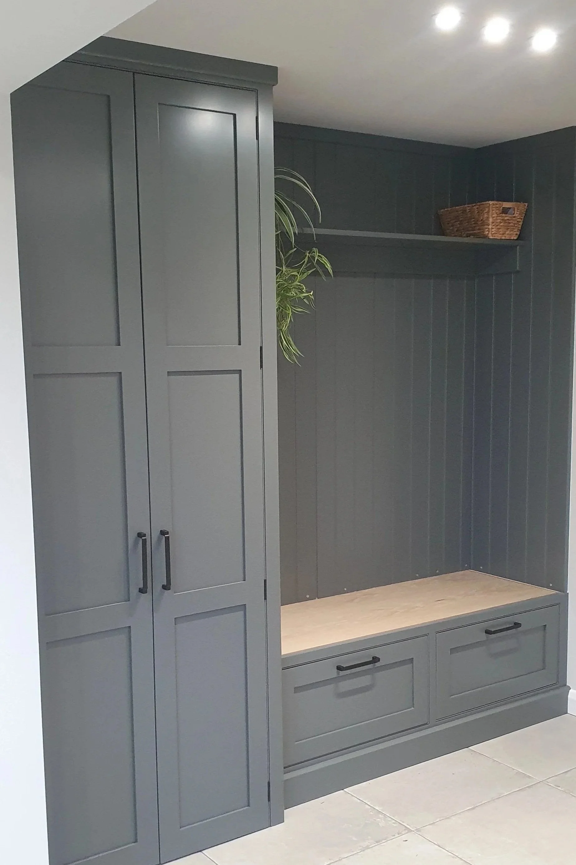 Gray built-in storage unit with tall cabinet, two drawers, a bench, a small shelf with a basket, and a potted plant in the corner.