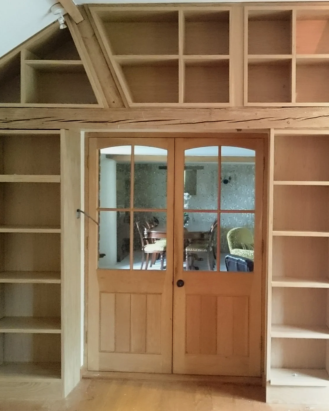 Wooden built-in shelves built into the angles of an Oak frame timber structure. And a pair of doors with glass panes leading to a dining room.