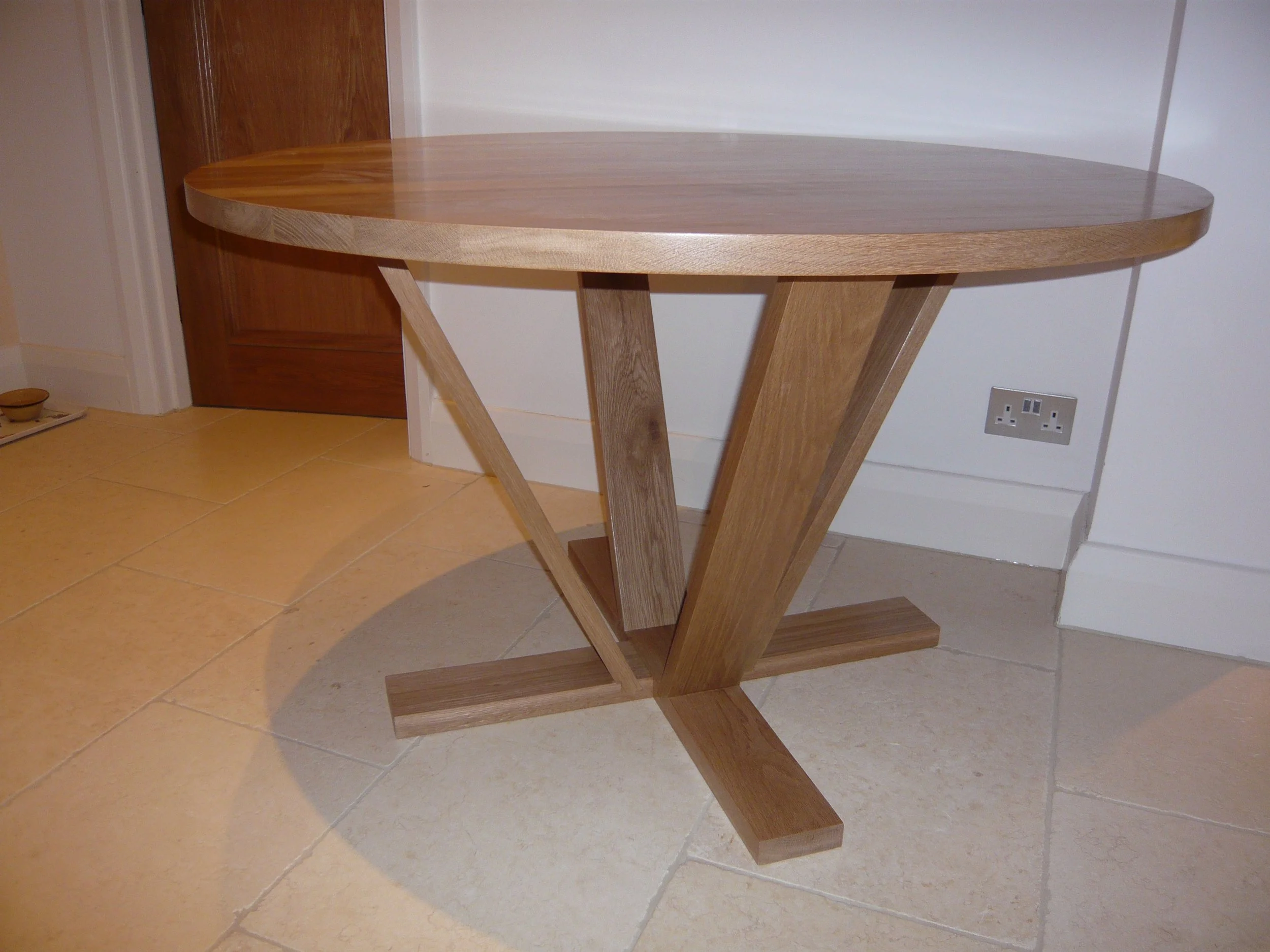 Wooden round-top table with angled support legs in a room with tiled floor and white walls.