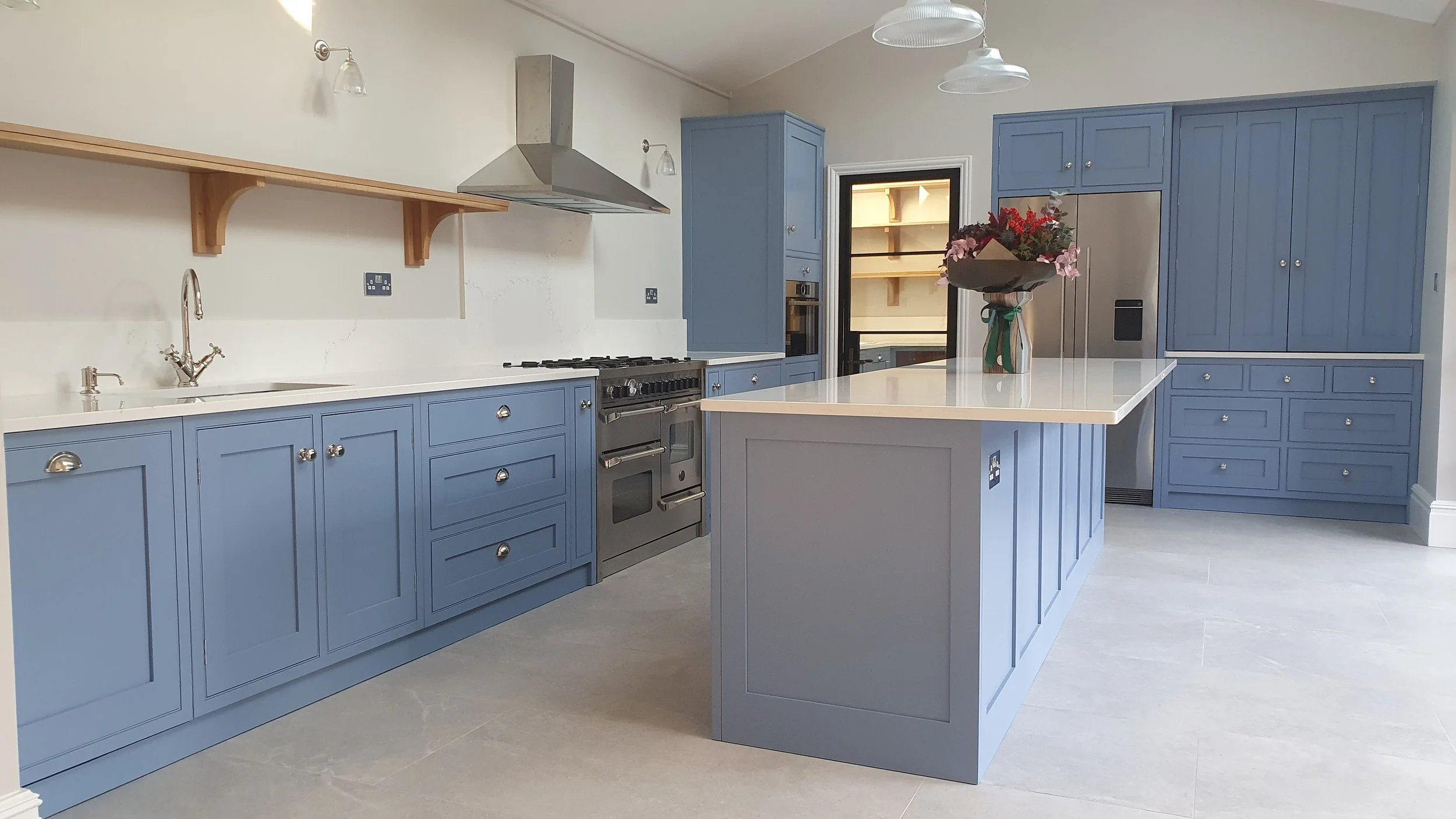 Modern kitchen with blue cabinets, white worktops and large island with seating. One Oak shelf on one wall.