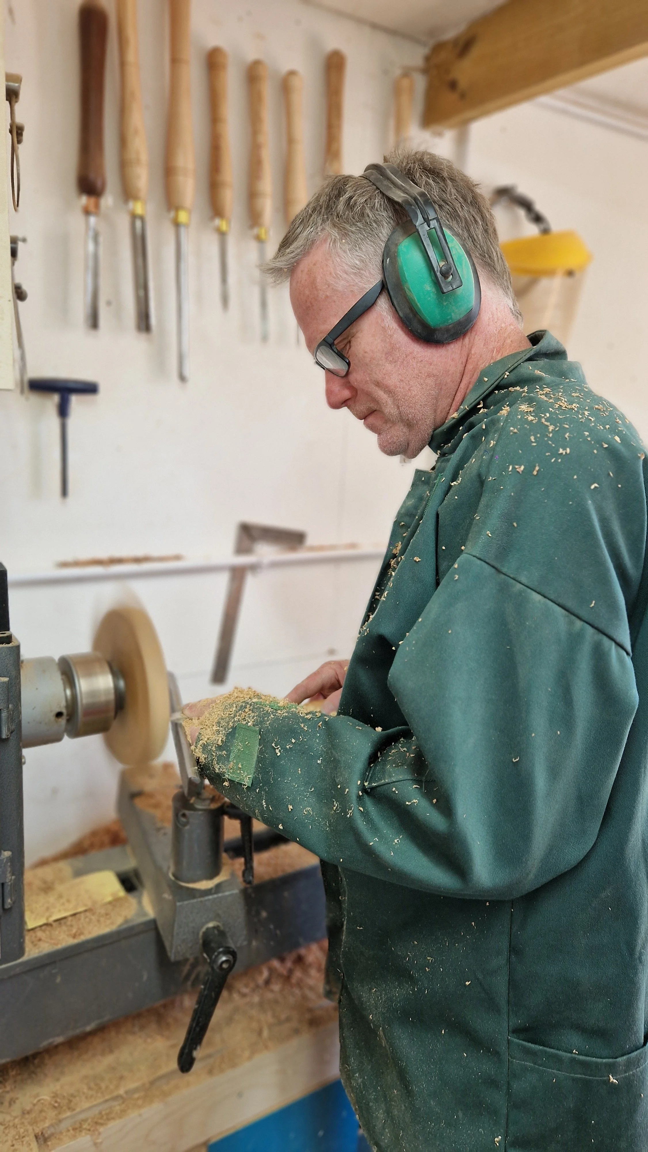 A man using a wood lathe with wood shavings on his green work jacket and headset, surrounded by woodworking tools on the wall.