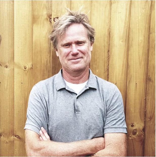 A middle-aged man with wavy blond hair and a slight smile, crossing his arms while standing in front of a wooden fence.