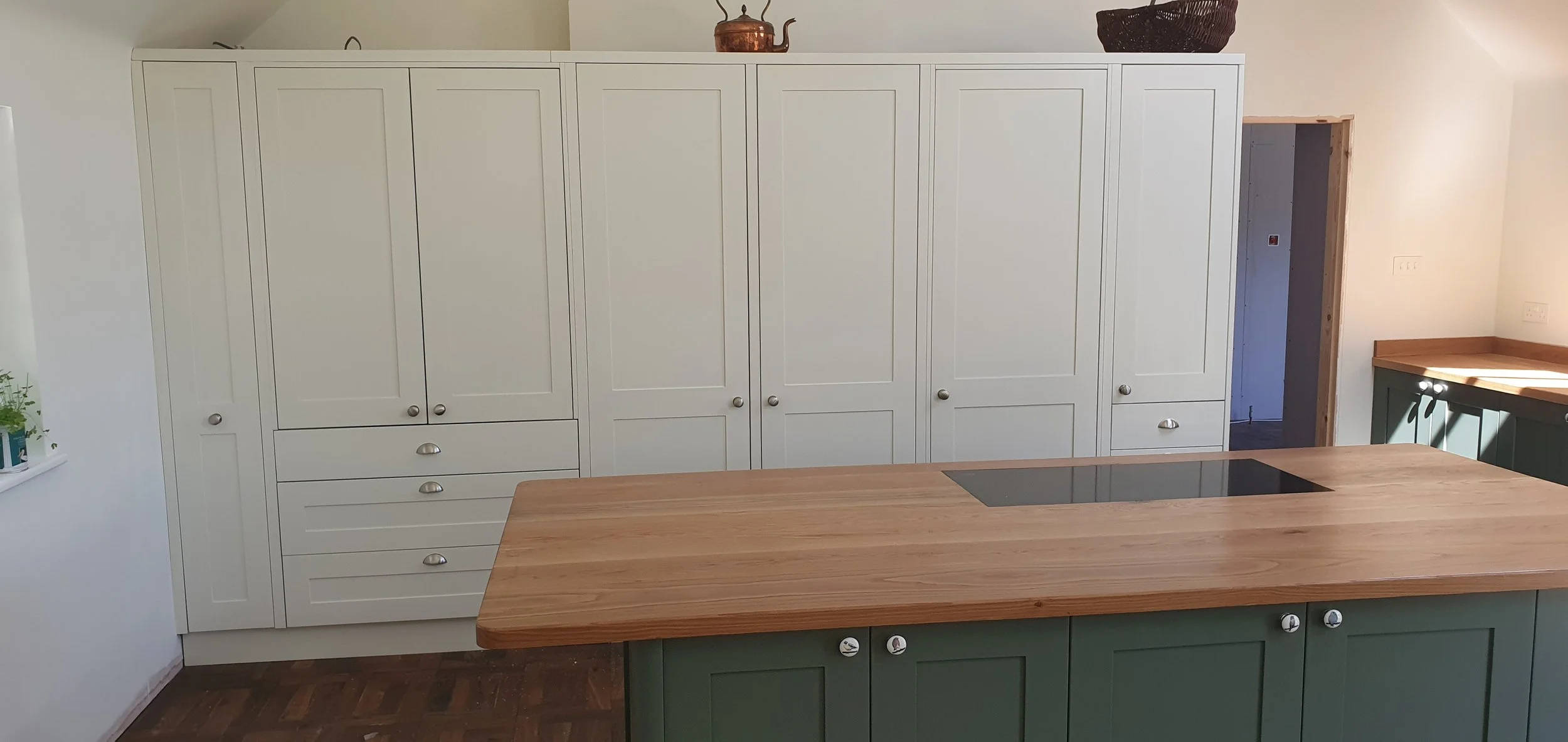 Large run of tall cream painted kitchen units that conceal the  oven and pantry cupboard with large green kitchen island 