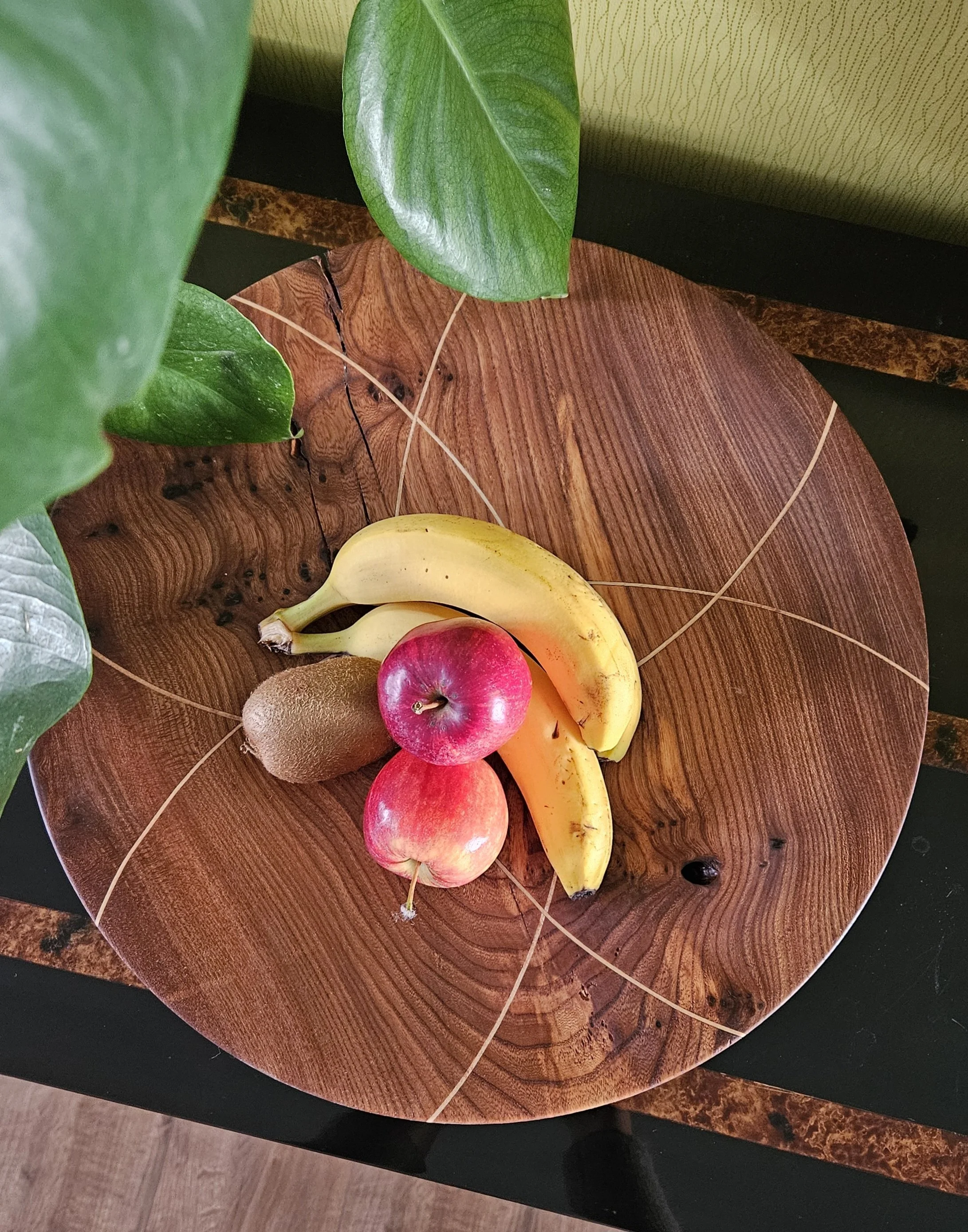 A wooden round tray with bananas, apples, a kiwi, and leaves on top.