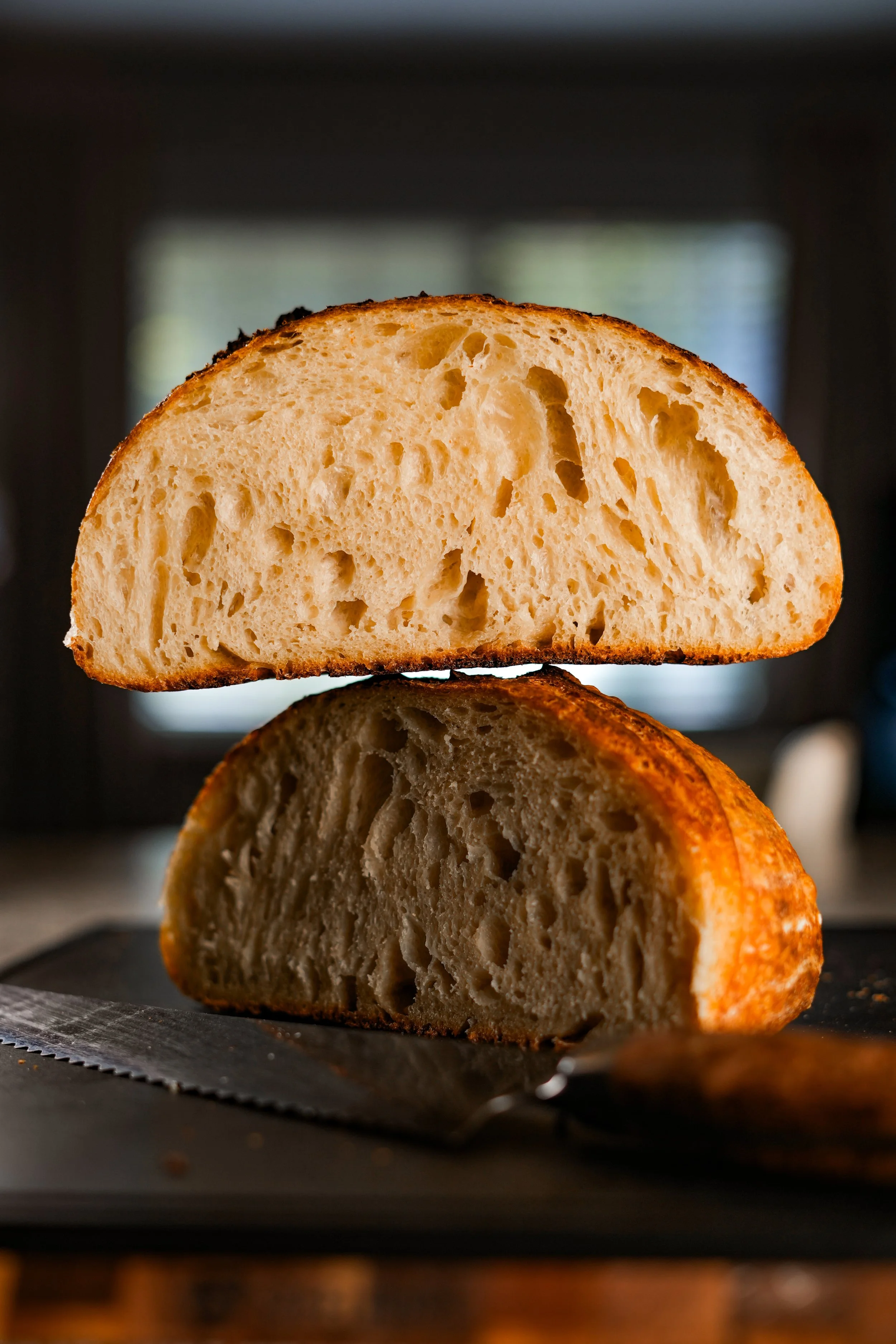A sliced loaf of artisan bread with a golden crust, placed on a dark cutting board with a bread knife nearby, with a blurred kitchen background.