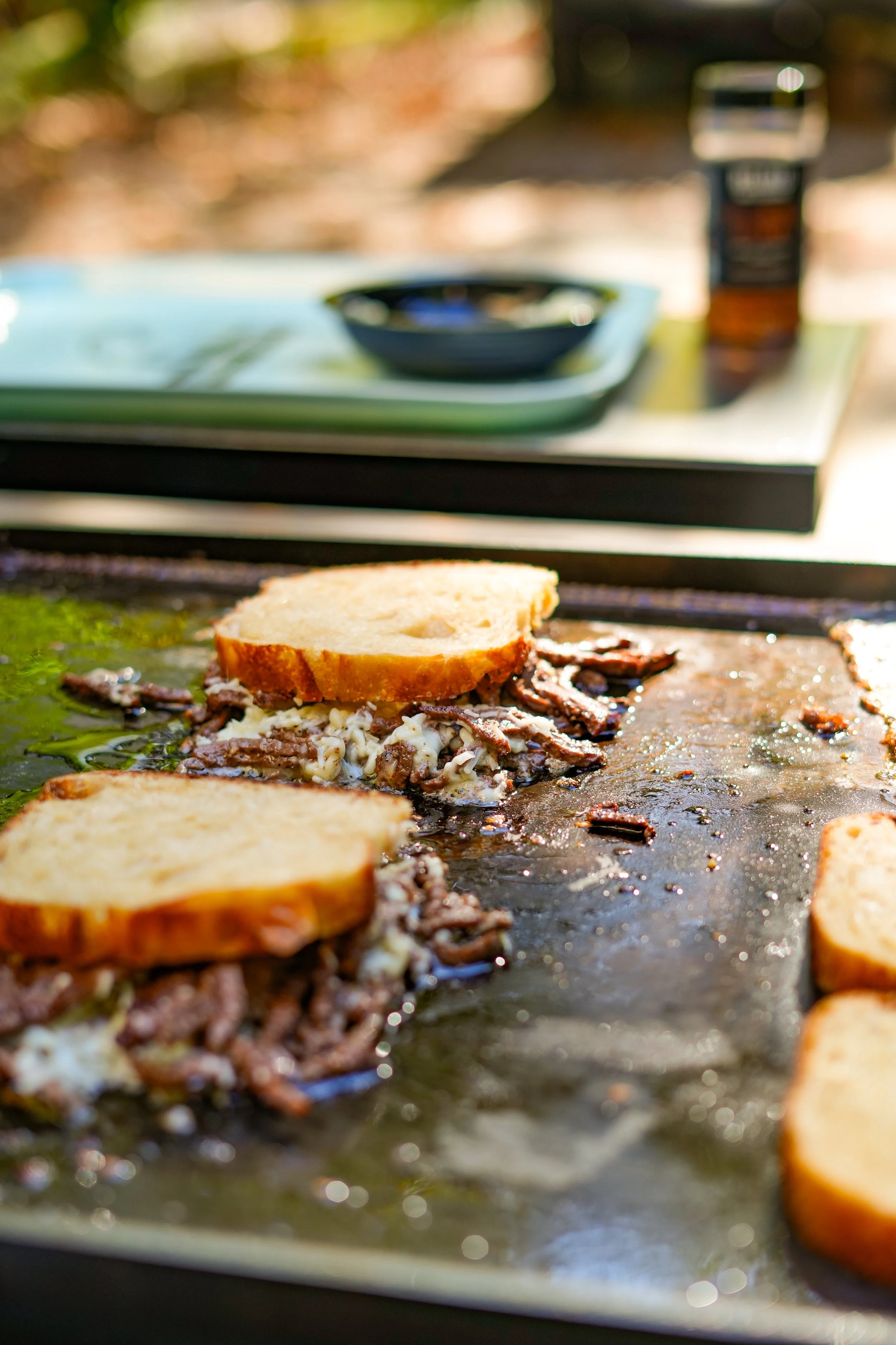 Close-up of grilled sandwiches with melted cheese, onions, and sliced bread on a griddle outdoors, with blurred placement of a bottle, black bowl, and tray in the background.