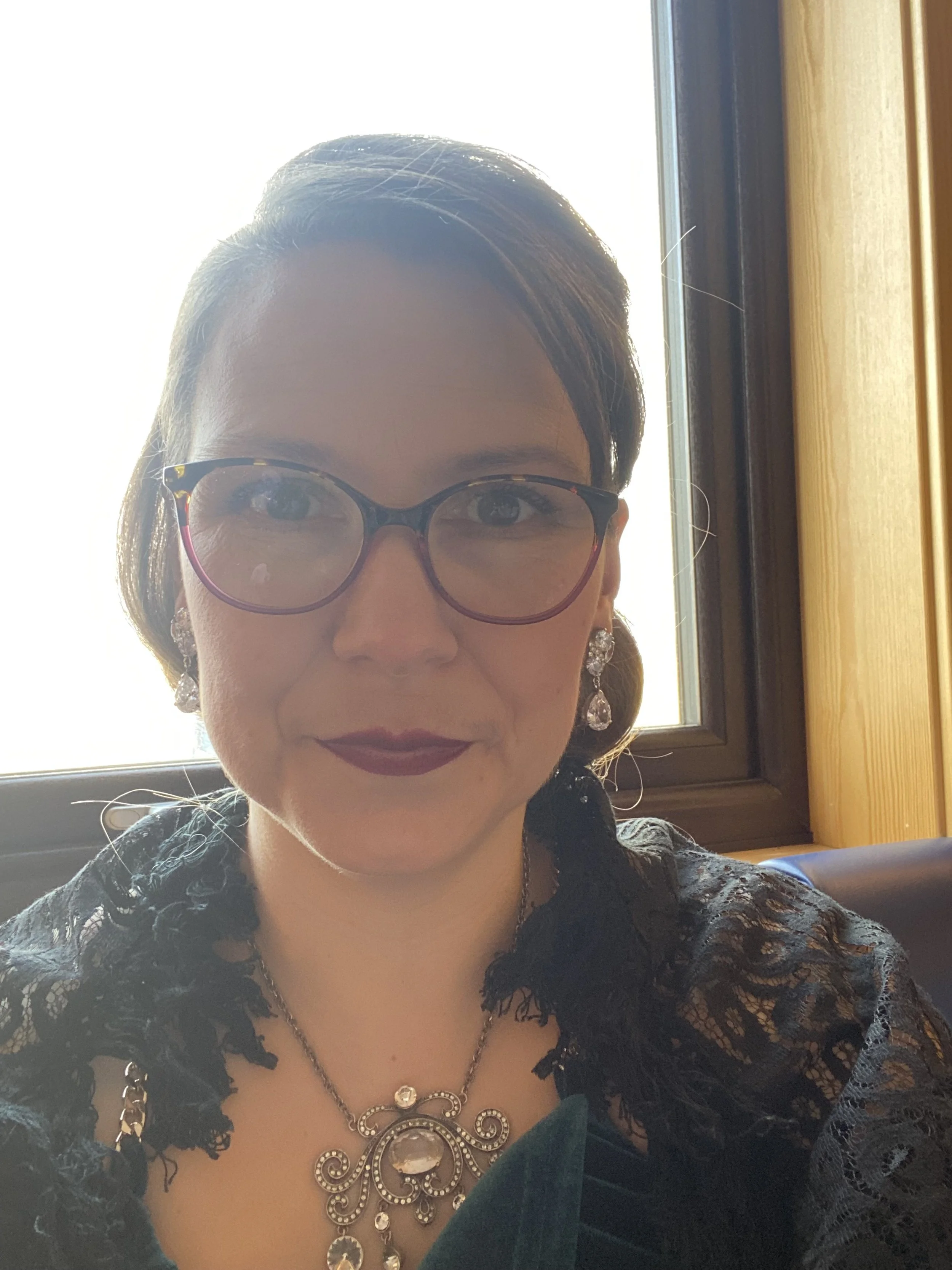 A woman with glasses and earrings sitting indoors near a window, wearing a black lace outfit and a ornate necklace.