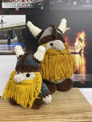Two handcrafted gnome plush toys with yellow beards, wearing blue and brown hats, placed on a wooden surface with a background of a poster showing a scene of a building fire near water.