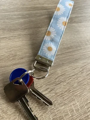 A keychain with a fabric strap featuring a daisies pattern, attached to keys and a small metal poker chip on a wooden surface.