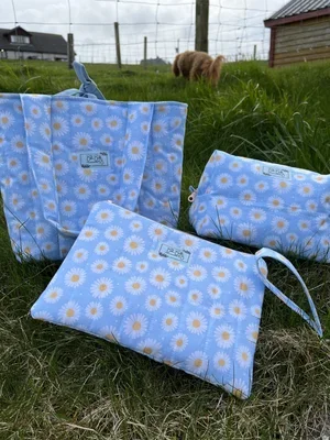 Three blue bags with daisy flower print, one handbag, one pouch, and one clutch, on grass with a cow and structures in the background.