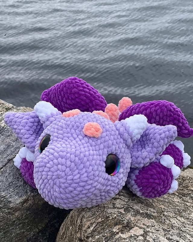 A plush purple and pink octopus toy with big eyes resting on a rock near water.