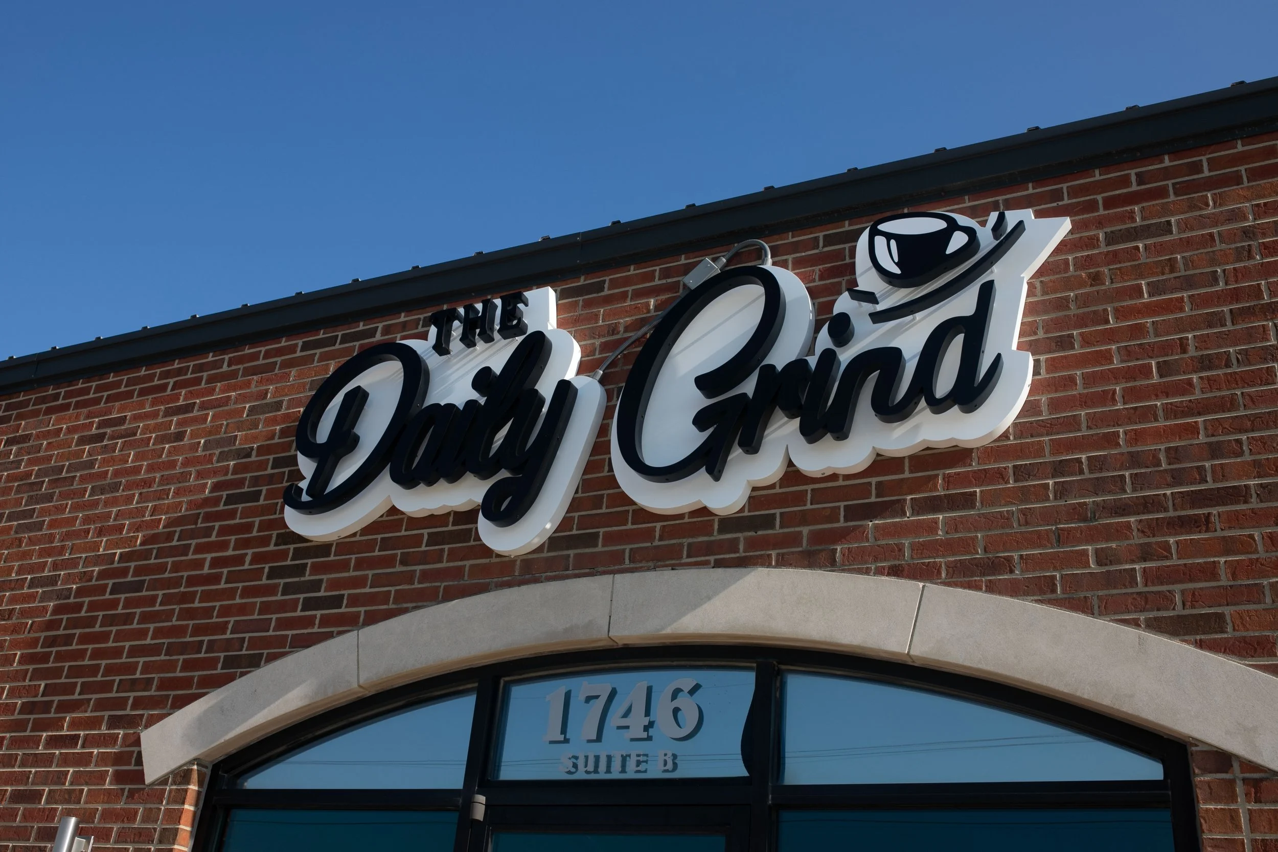Sign for The Daily Grind coffee shop on a brick building with the street address 1746 Suite B below the sign.