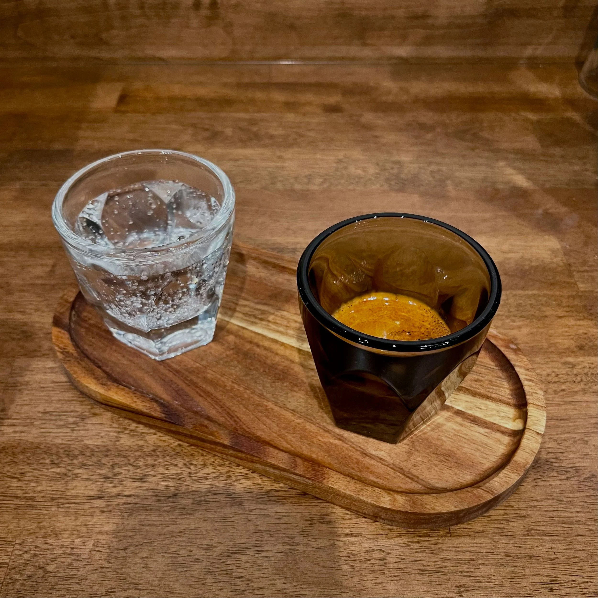 A glass of water with ice cubes and a small cup of espresso on a wooden tray placed on a wooden table.