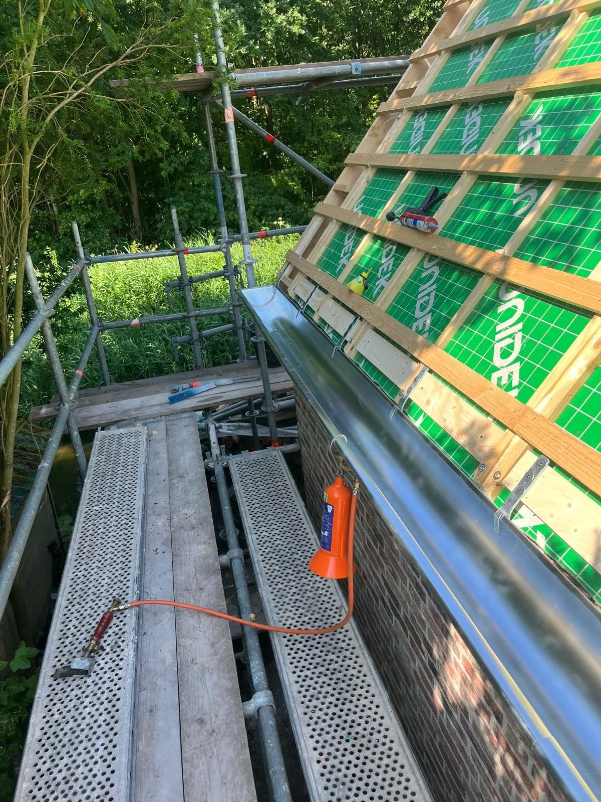 Bouwsteiger op een dak met houten balken en groene isolatiepanelen, omgeven door groen bomen en struiken.