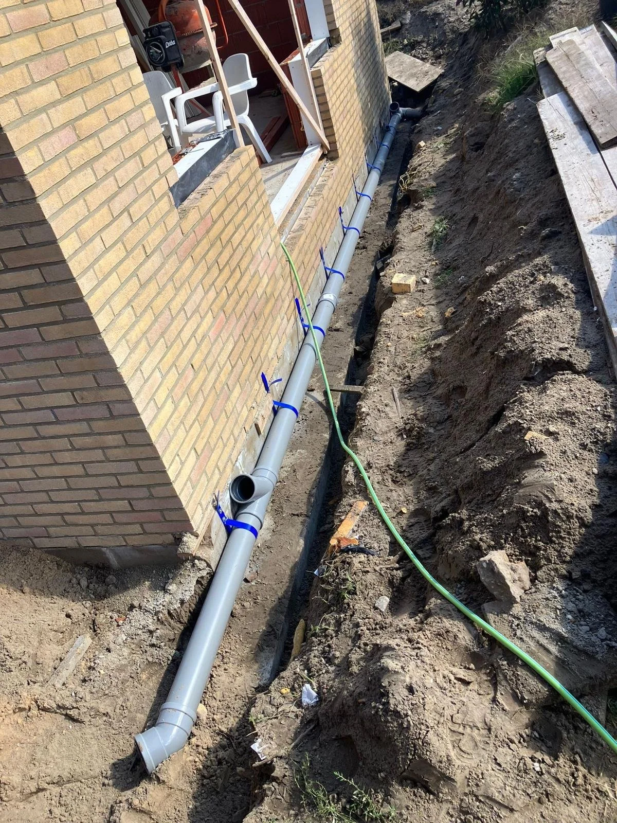 Installatie van drainagesysteem langs de fundering van een huis met bakstenen muren, met grote buizen en een groen snoer op een bouwterrein.