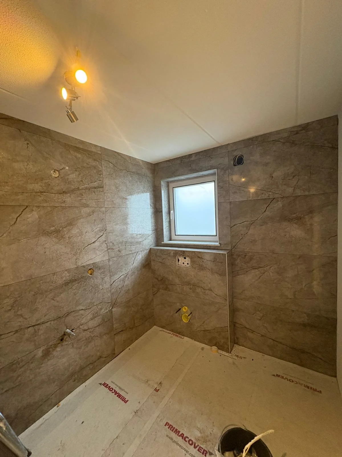 Een kamer onder nieuwbouw met beige marmeren wandtegels, een klein raam, ingebouwde elektrische aansluitingen en een vloer met isolatiemateriaal. Er zijn geen meubels of andere voorwerpen.