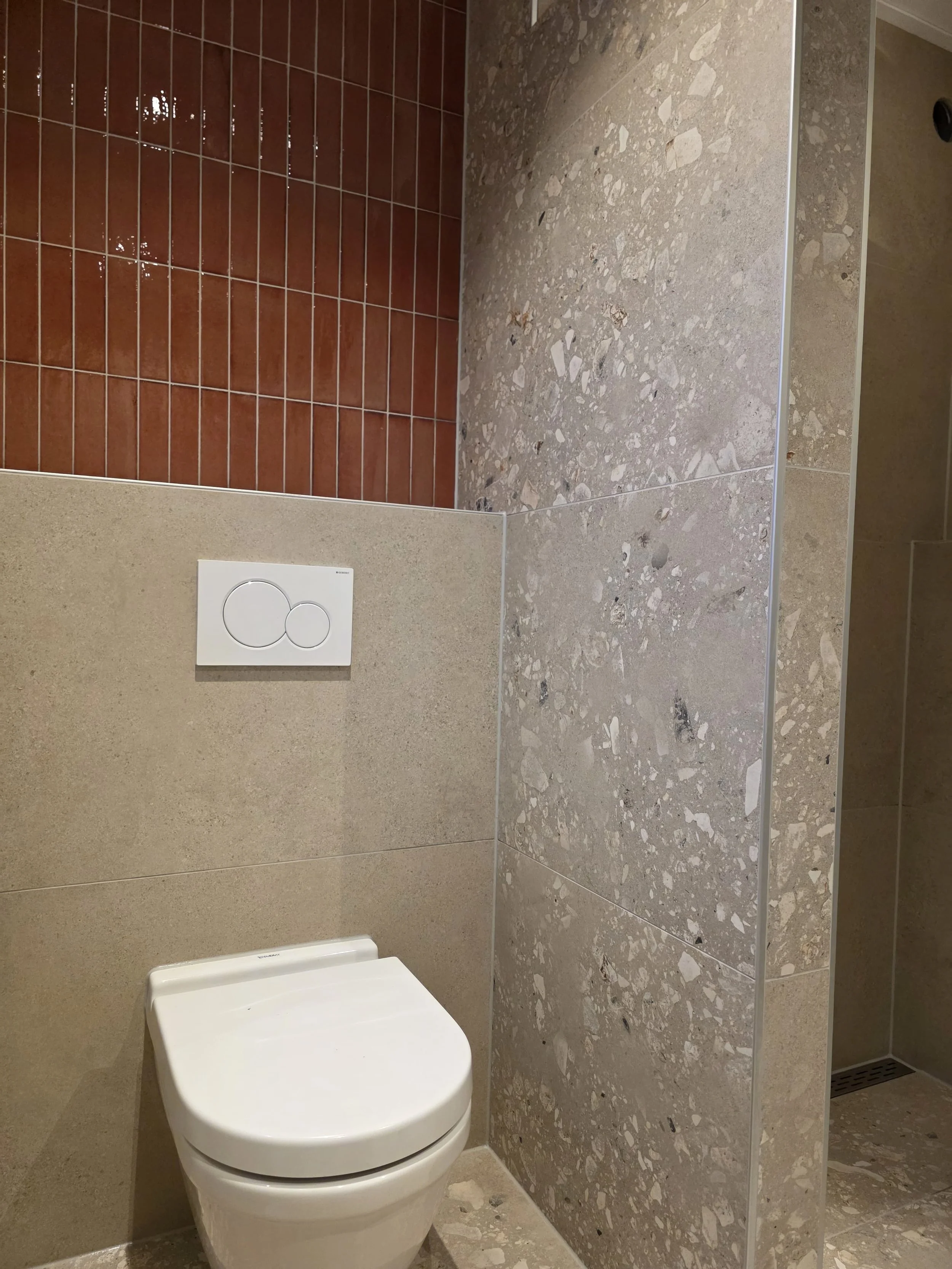 Een modern toilet met een inbouwreservoir en een dubbele bedieningsknop, gelegen in een badkamer met tegels in beige, rood en terrazzo stijl.