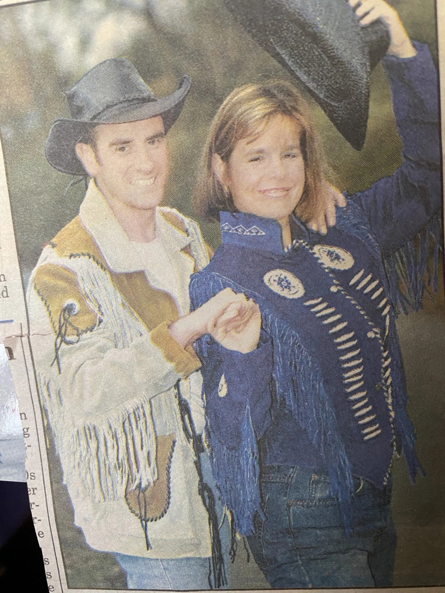 Two women smiling, one wearing a texas cowboy hat and western attire, the other in a blue button-up shirt with fringe and embroidery, both holding cowboy hats.