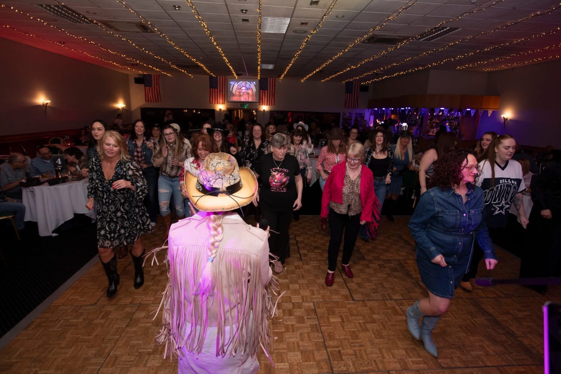 People line dance at a country-themed event in a decorated banquet hall with American flags and string lights.